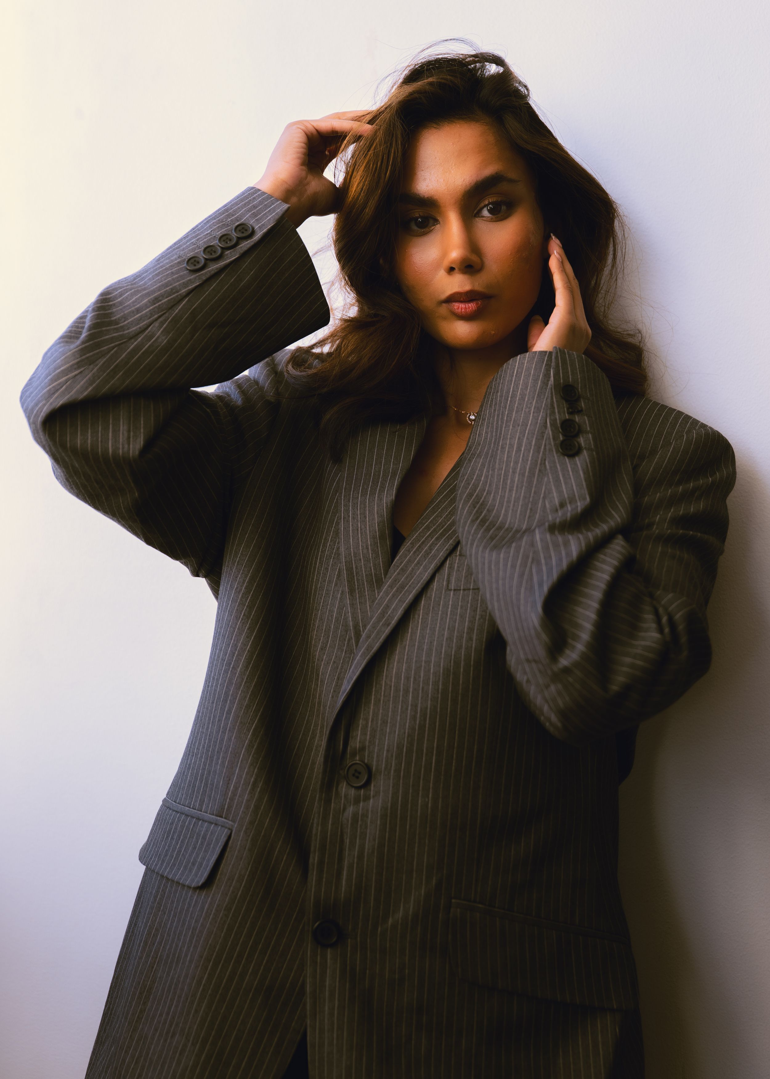 Stylish woman in oversized grey pinstripe suit poses with hands in hair against white background.