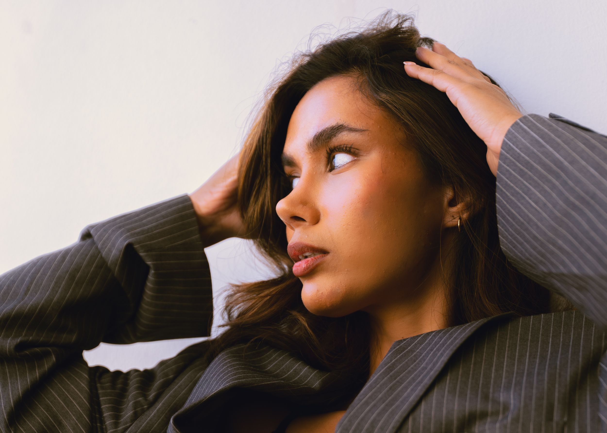 Young woman in grey pinstripe blazer poses thoughtfully with hand in hair against white wall.