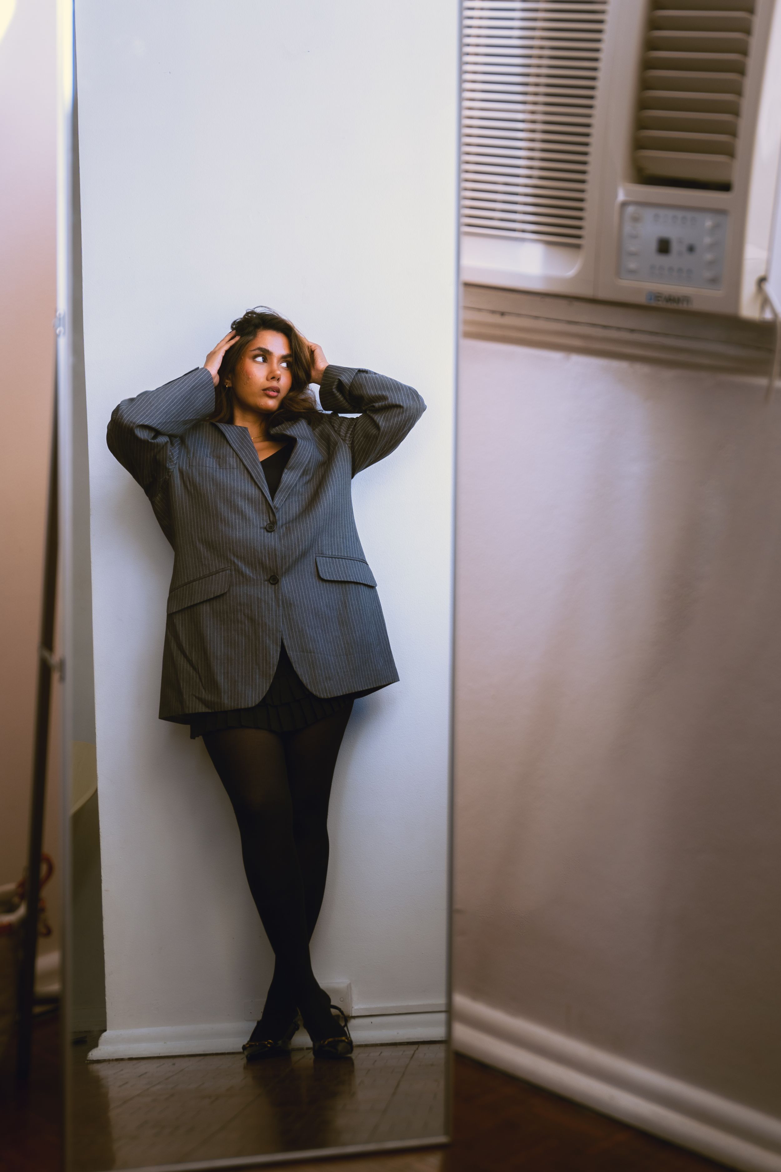 Woman in grey blazer and black tights leans against white wall in dimly lit room.