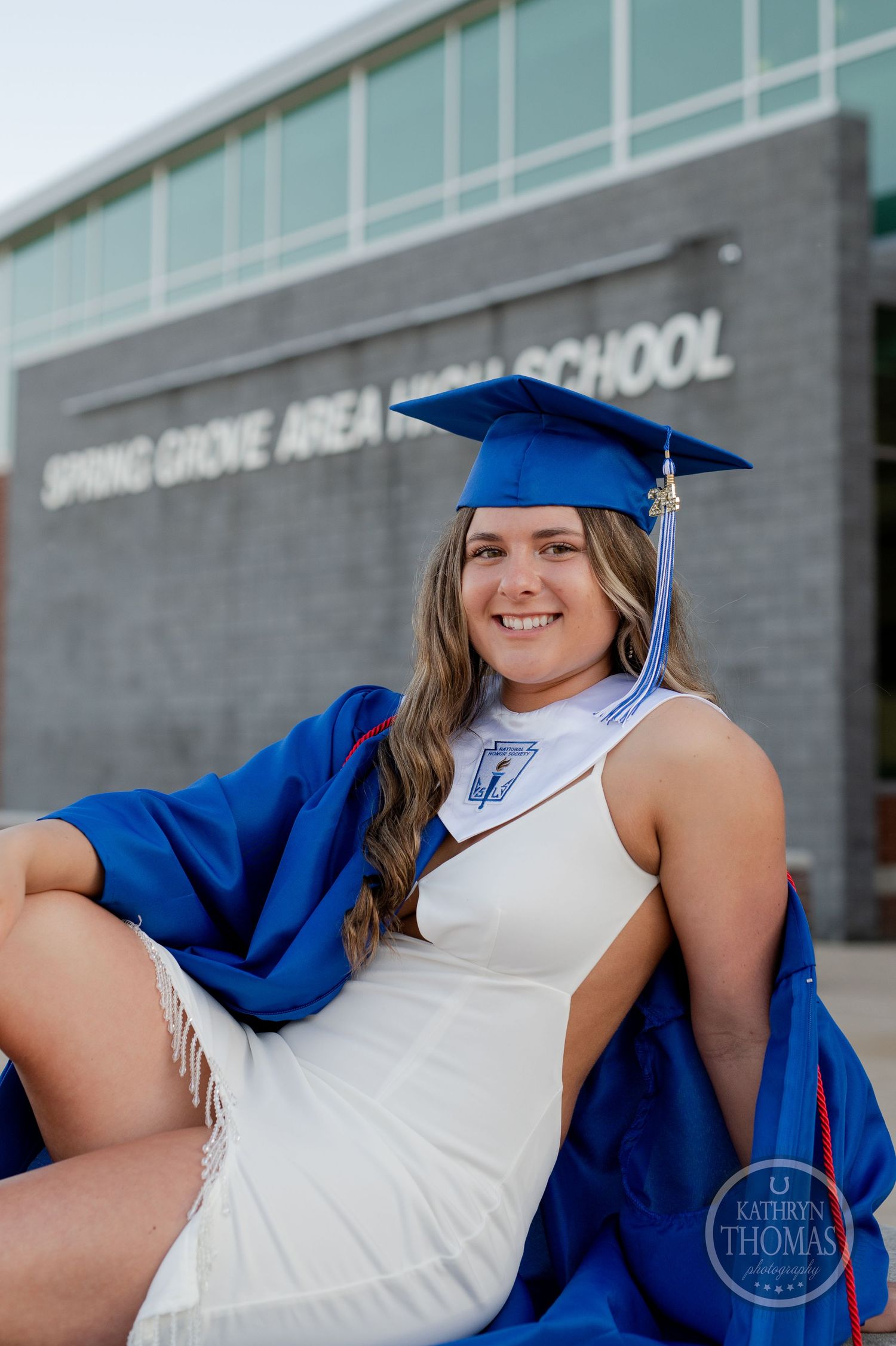 Graduate in white dress and blue regalia poses by Spring Grove Area High School entrance.
