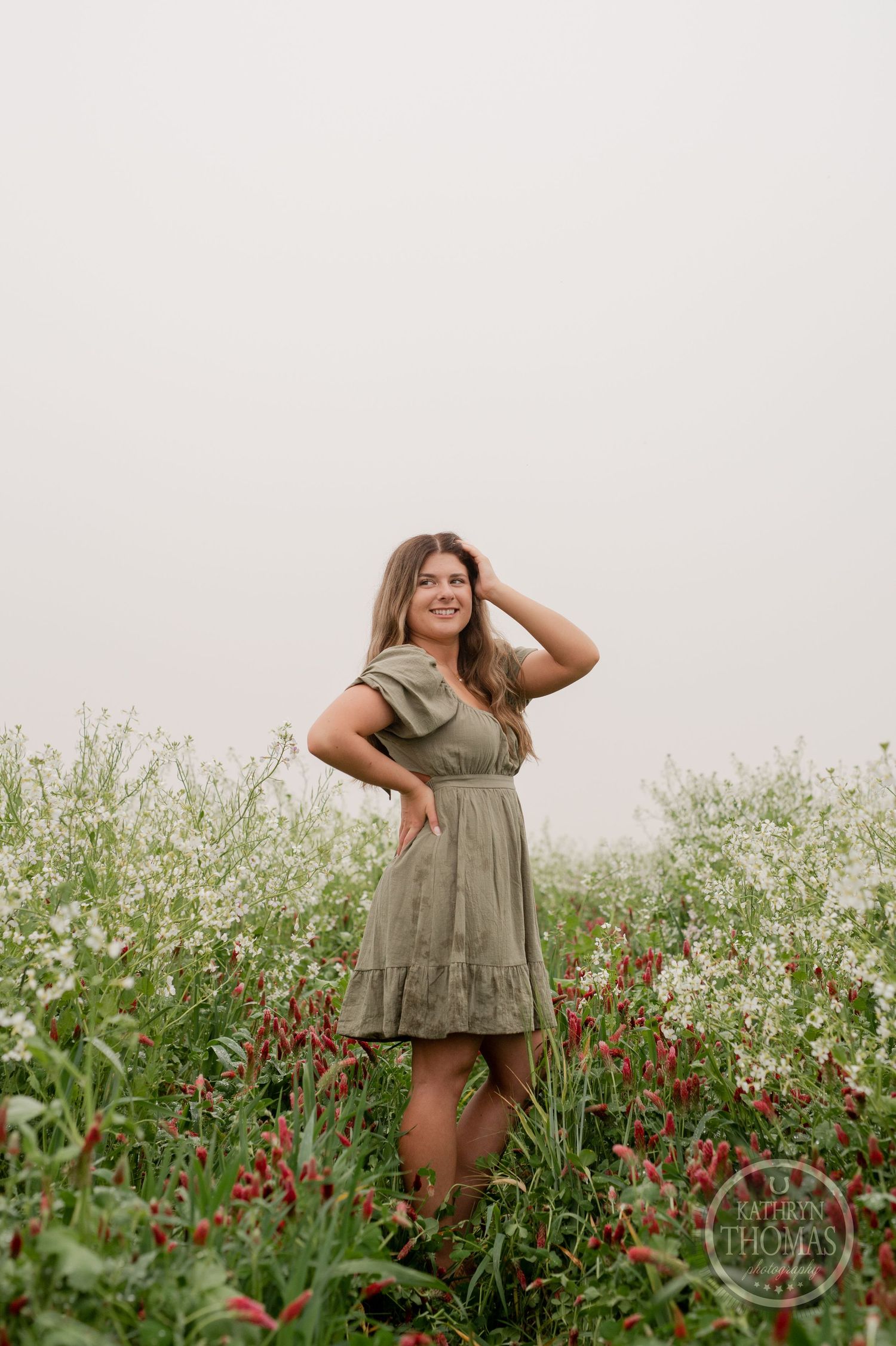Model in green dress poses naturally among wildflowers in a misty field setting.