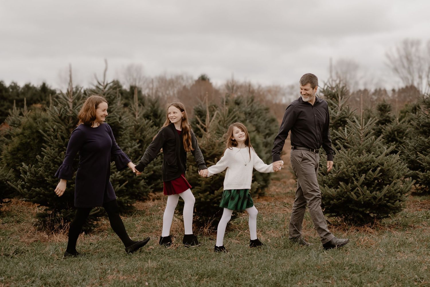 Family holding hands and walking together through rows of evergreen trees at a Christmas tree farm on an overcast day.