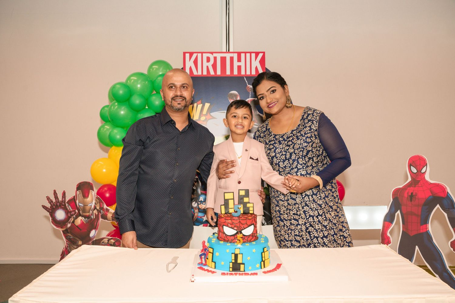 A birthday celebration features a superhero-themed cake with Spider-Man and Iron Man decorations and green balloons.