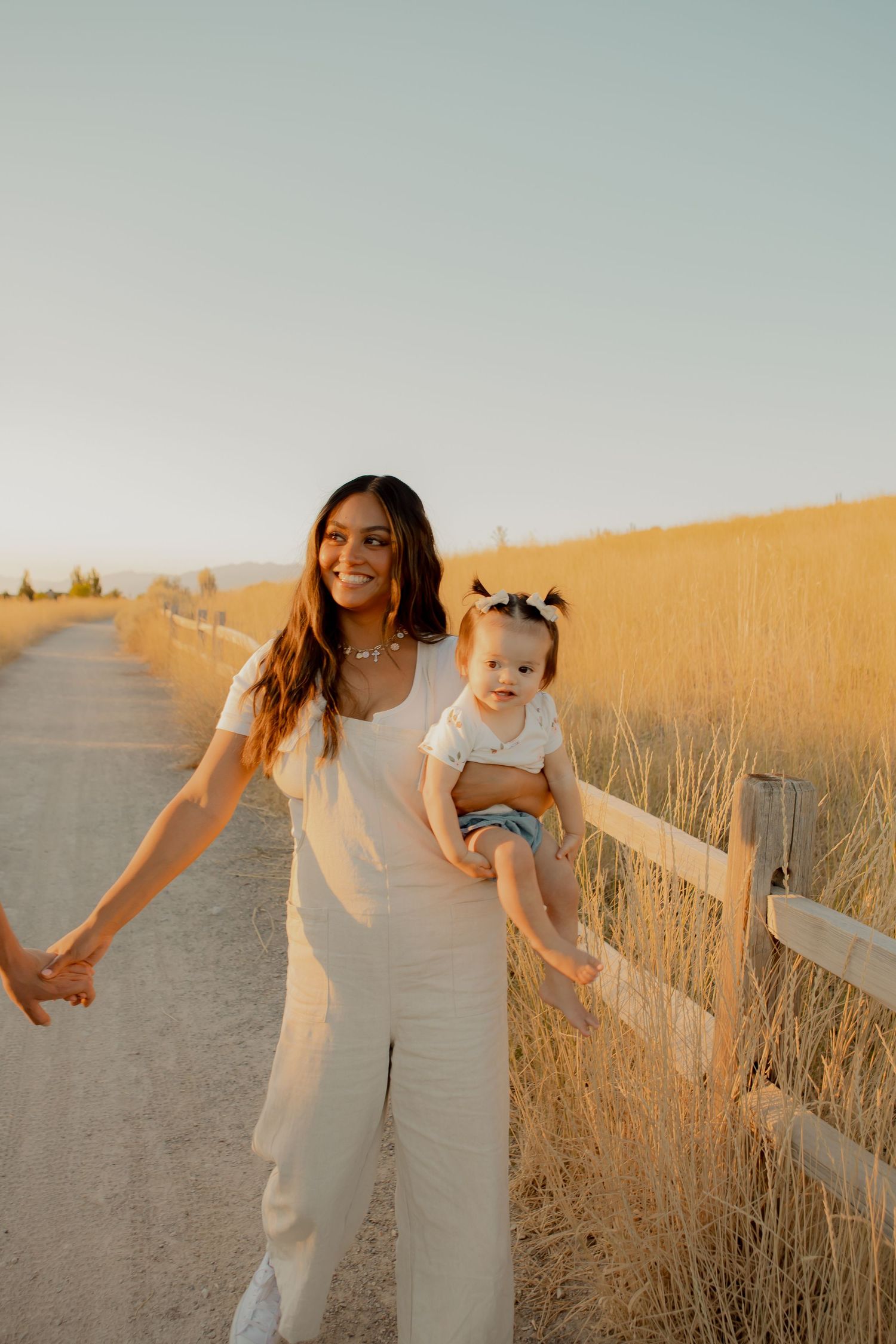 Woman in white outfit holding a toddler along a rural fence-lined path at golden hour.