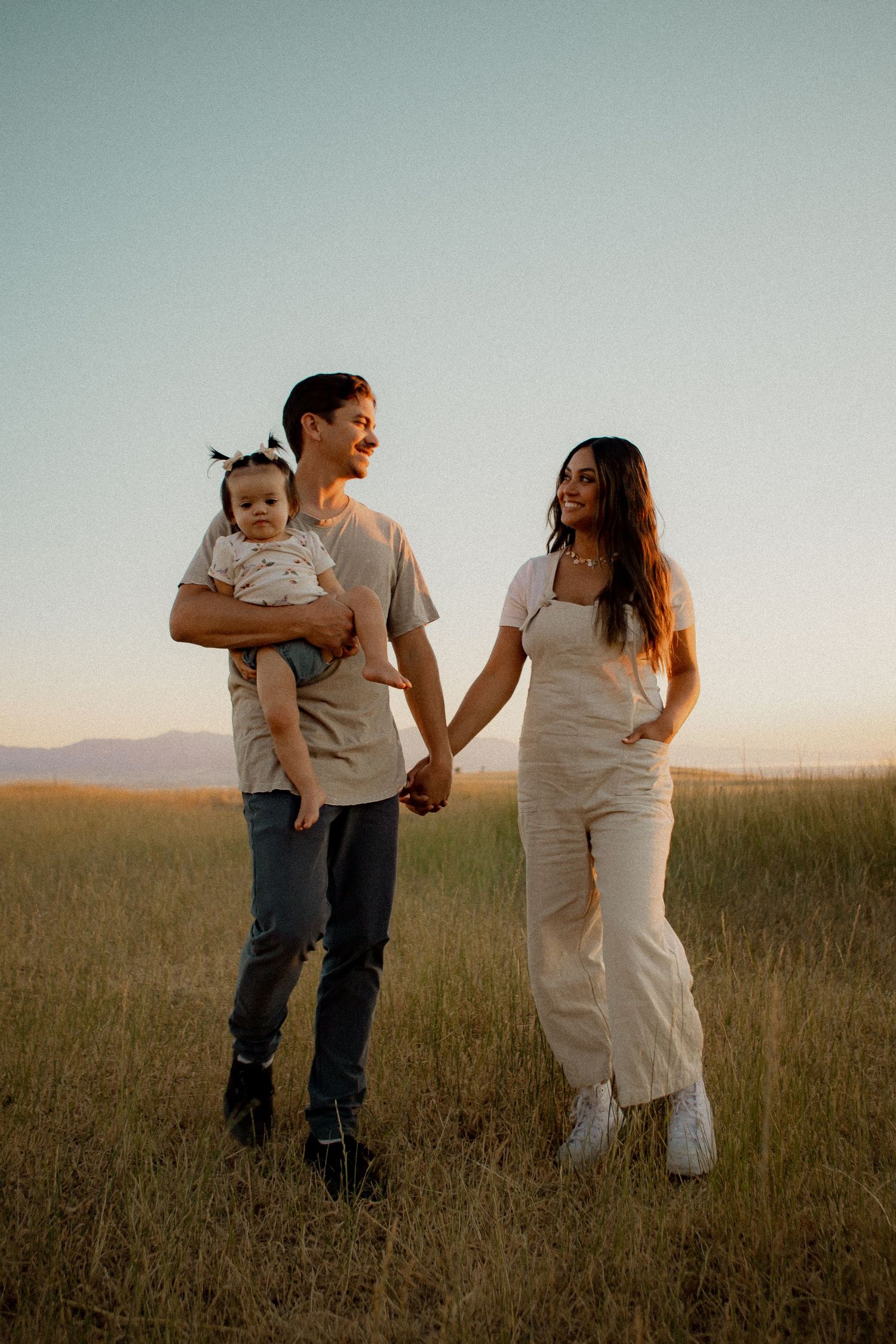 Family walking together in a golden field at sunset, holding hands and carrying a small child.