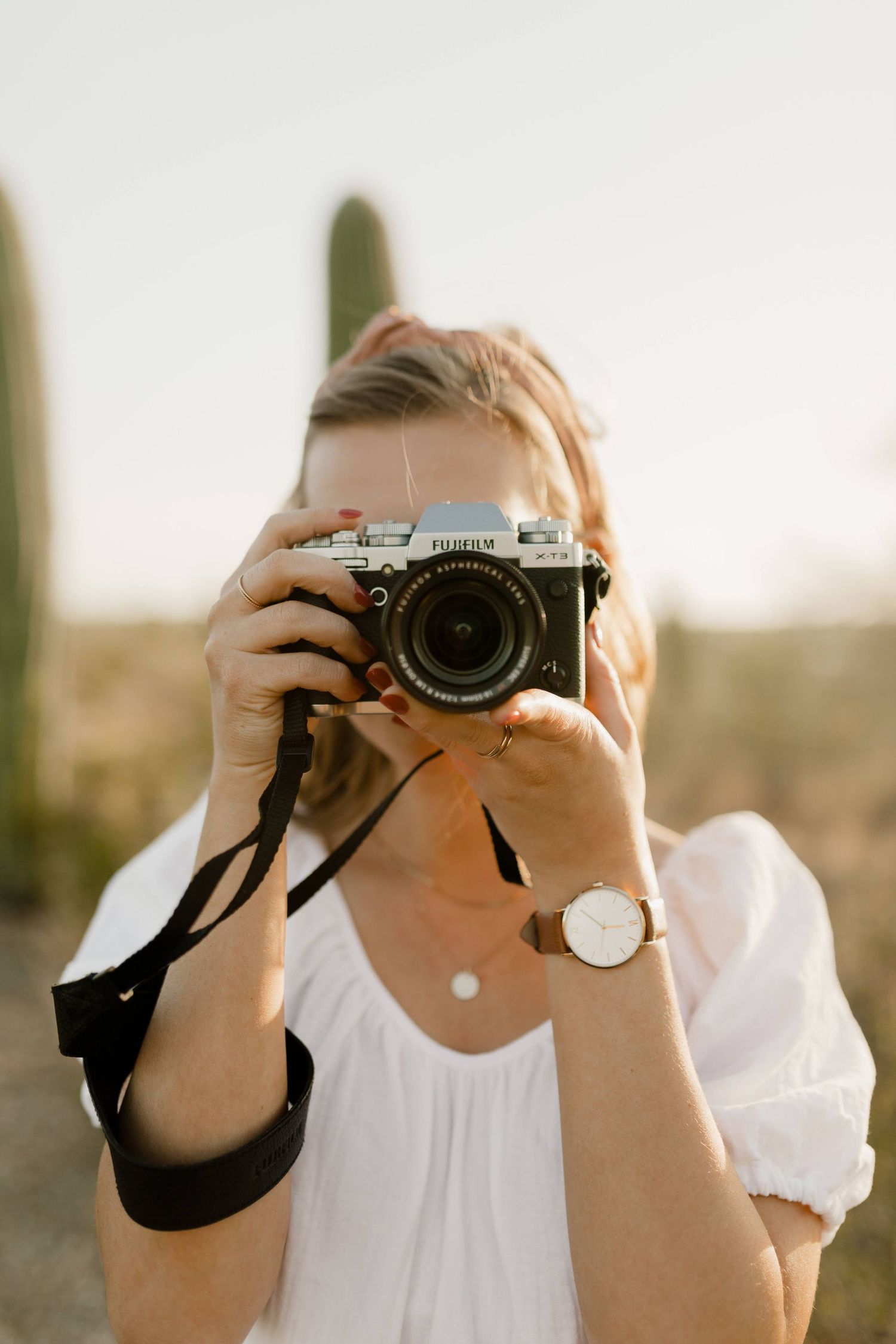 Photographer in white blouse holding vintage camera with black strap against blurred outdoor background at sunset.