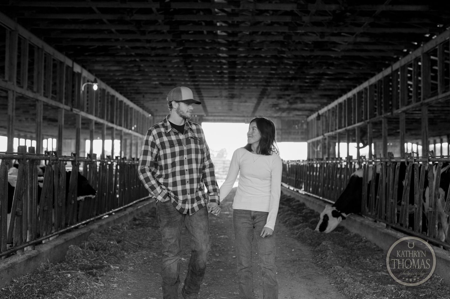 A black and white photo of a couple walking through a barn with cattle stalls.