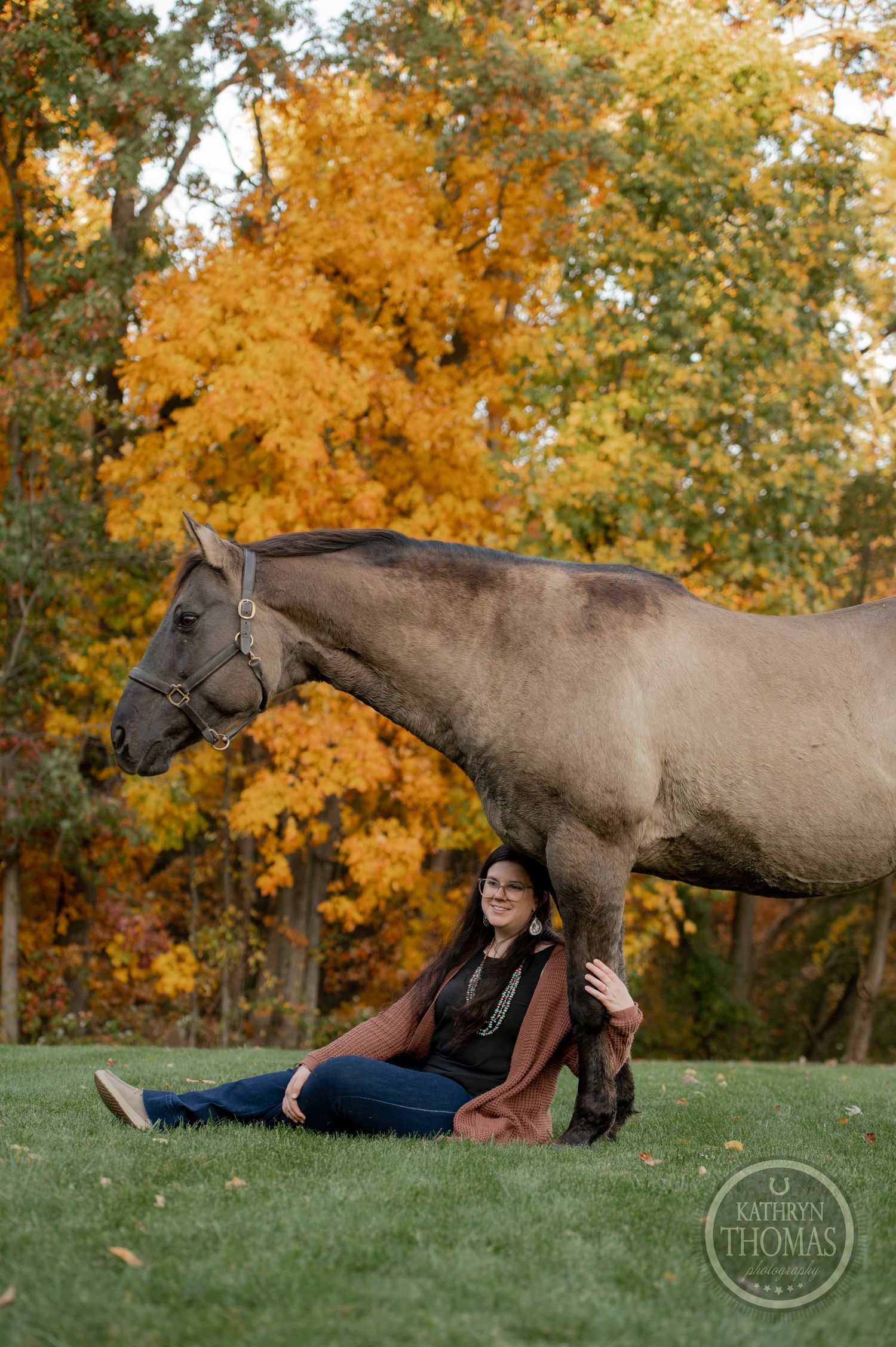 A gray horse stands next to someone relaxing on grass with golden fall trees in the background.