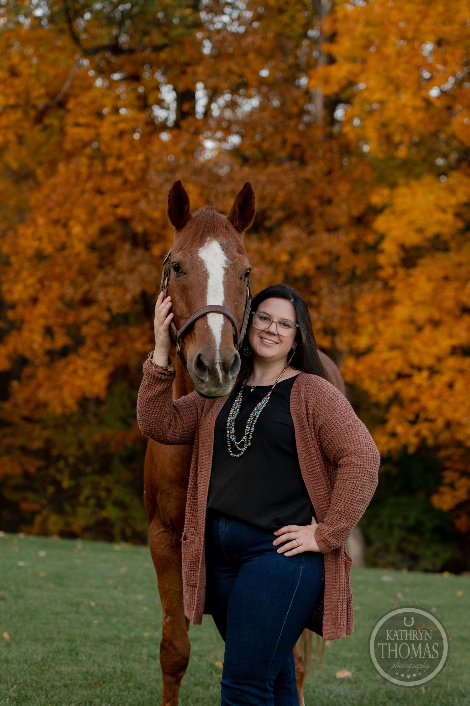 A chestnut horse with white blaze stands proudly next to someone amid fall foliage.