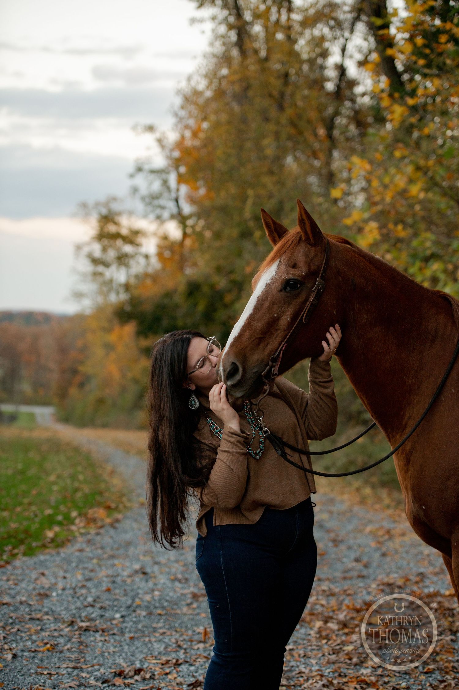 A woman in a brown sweater affectionately pets her horse on a fall trail lined with colorful trees.