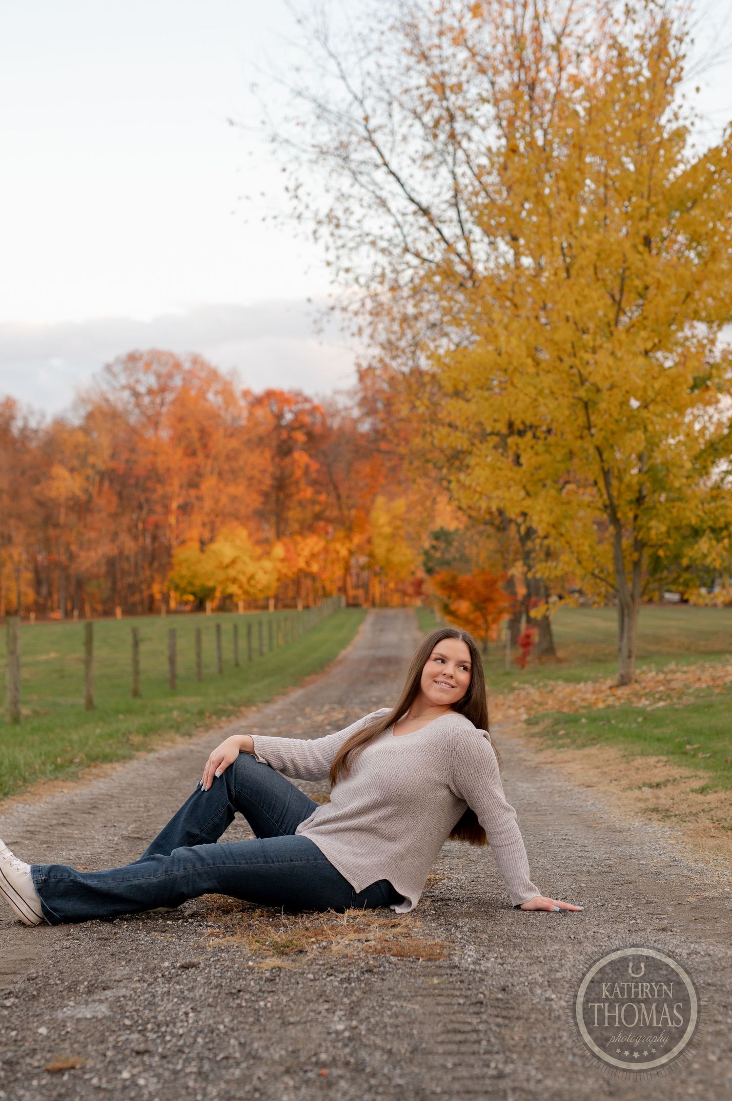 Person reclines on dirt path with autumn trees and fence in rural setting.