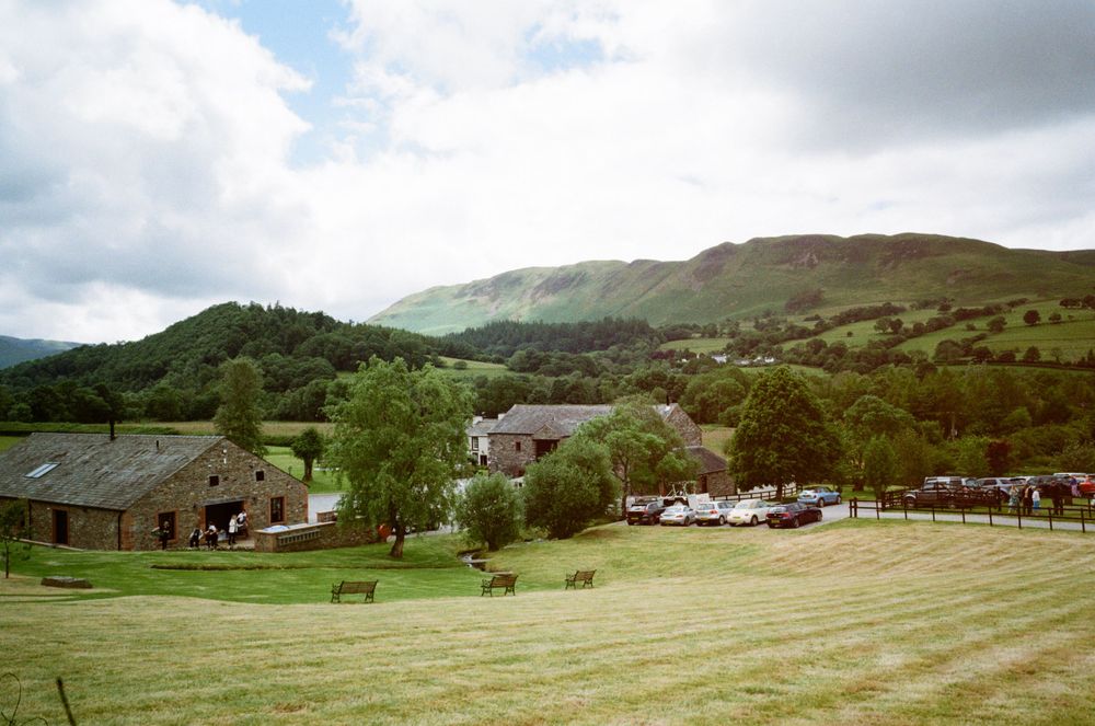 Scenic rural farm with stone buildings nestled in a green valley surrounded by wooded hills under a cloudy sky.