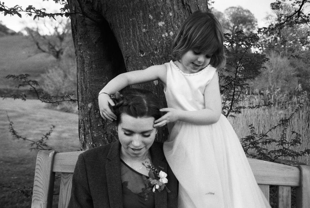 Black and white photo of a young flower girl playfully styling a seated womans hair by a tree.
