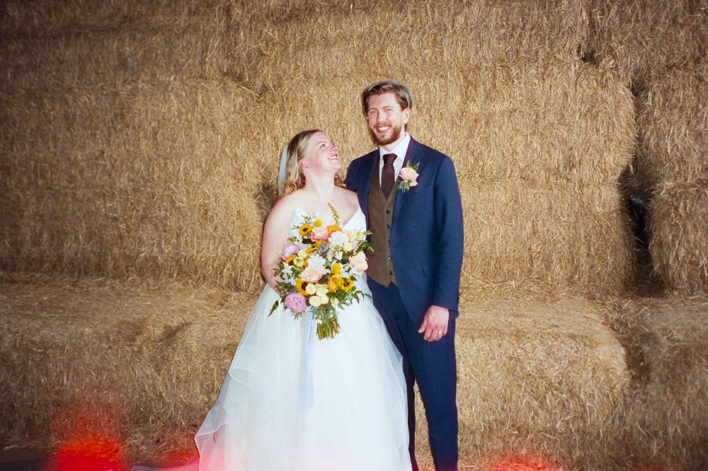 Bride and groom pose in front of hay bales in a rustic barn, bride holding a colorful bouquet.