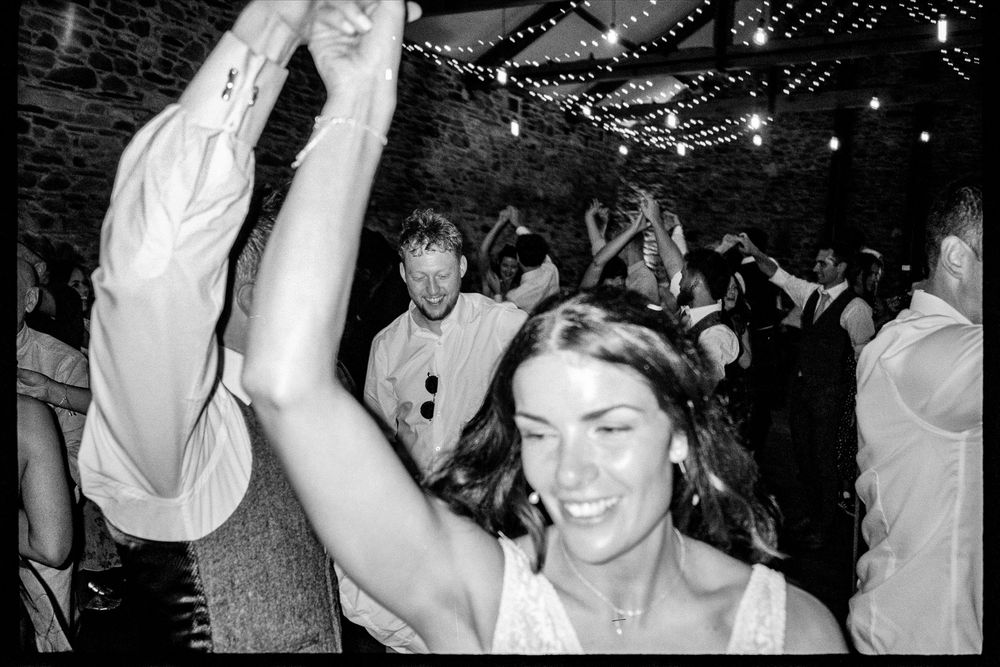 Smiling bride dancing with arm raised among wedding guests in black and white photo at barn reception.