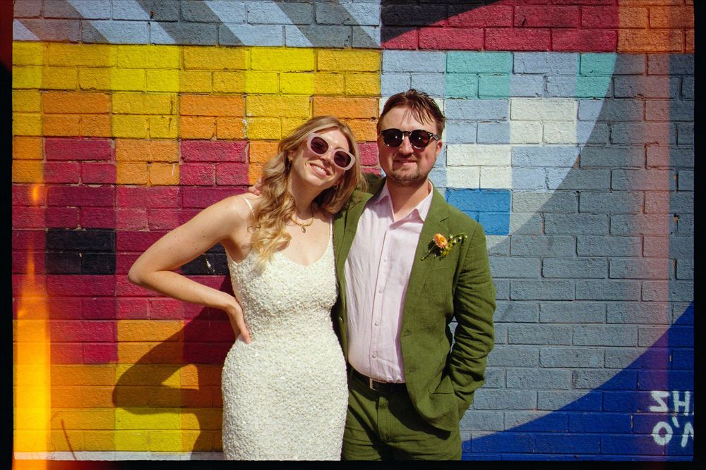 Couple posing in front of colorful graffiti mural wall, bride in white lace dress and groom in green suit wearing sunglasses.