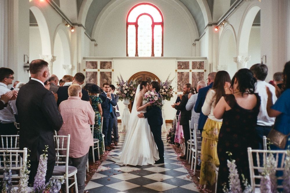 Bride and groom share first kiss at church wedding ceremony aisle with guests standing and floral decorations.