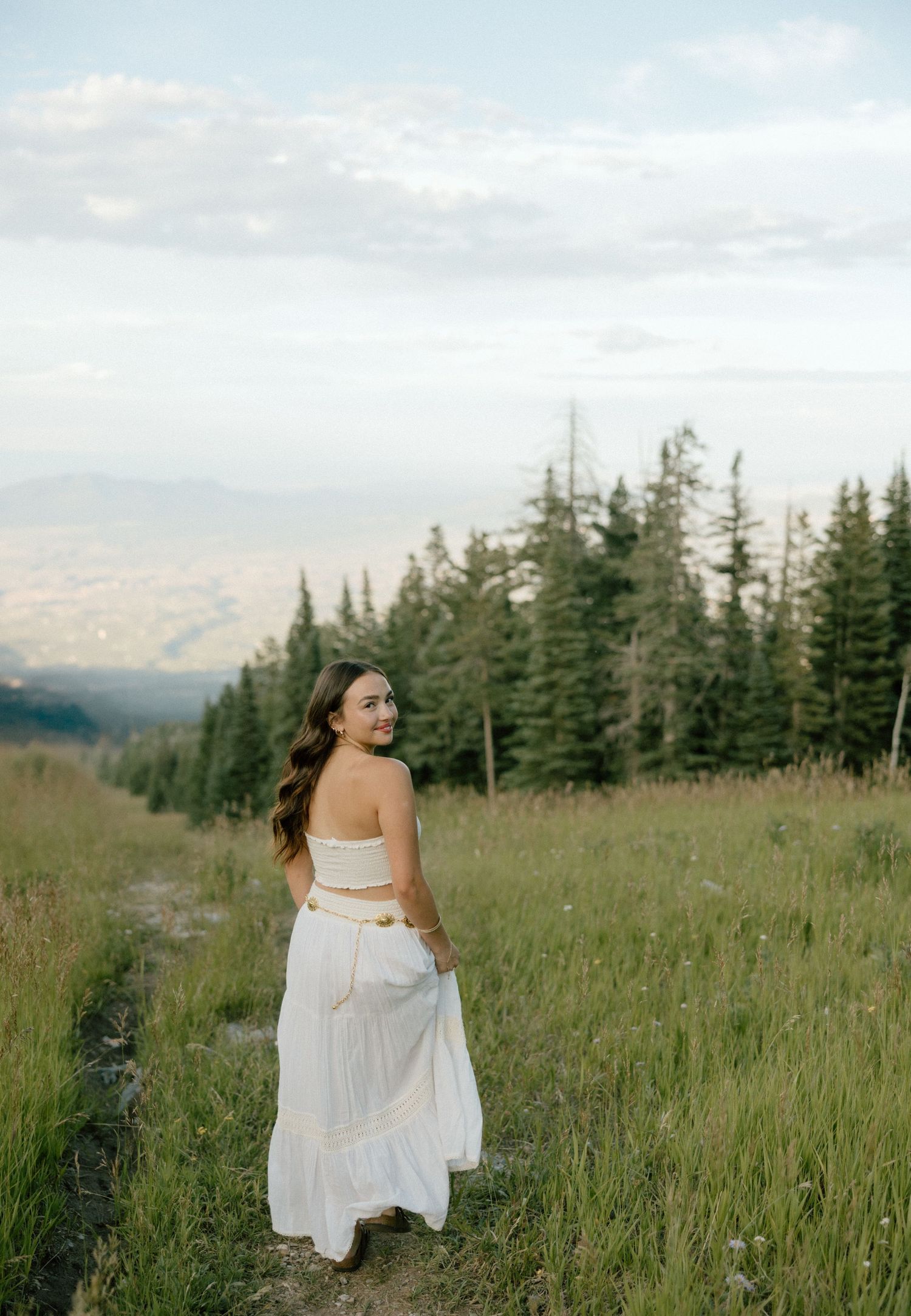 A woman in a flowing white dress twirls gracefully through a mountain meadow surrounded by evergreen trees.