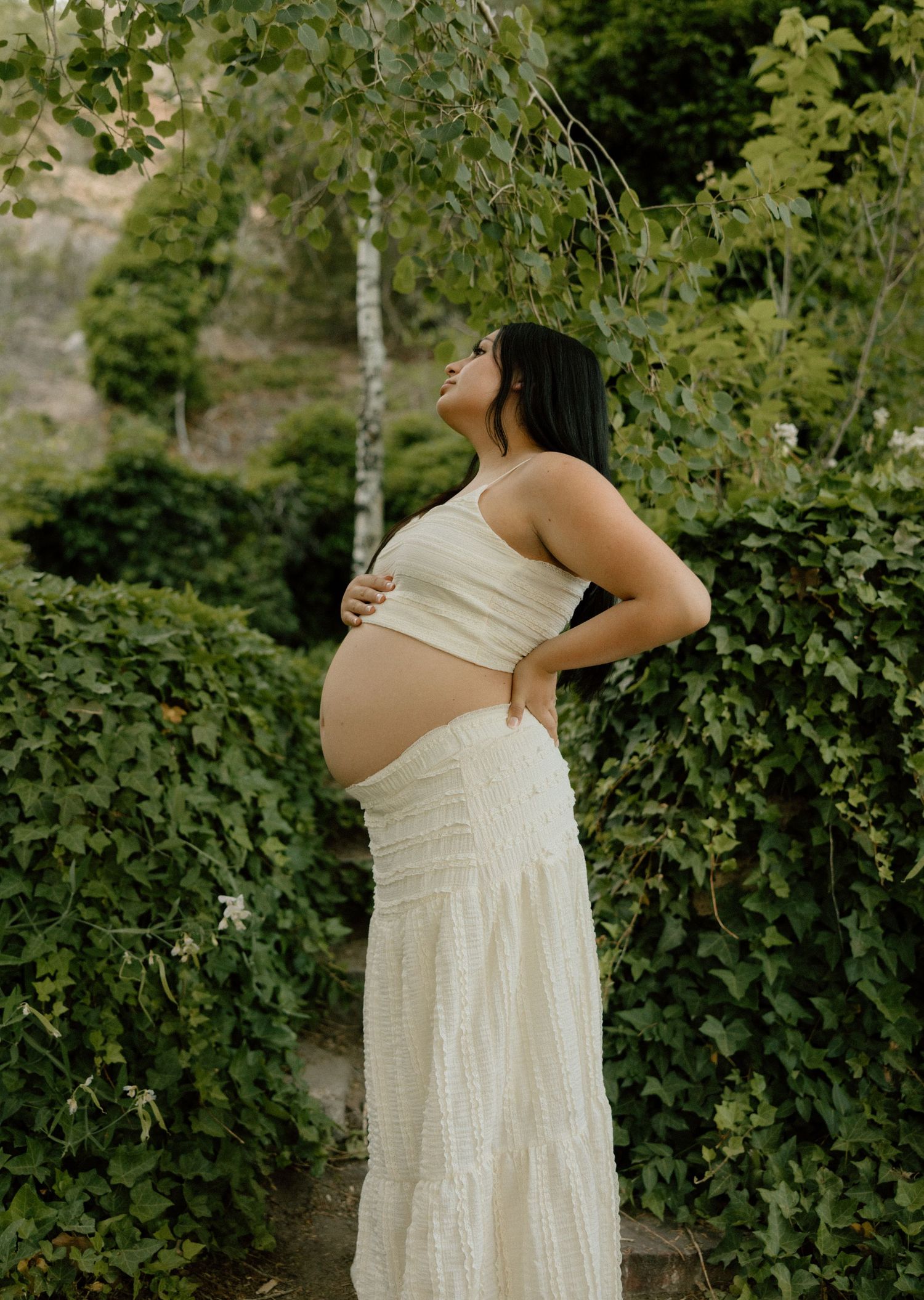 A cinematic photo of an expecting mother in a flowing white dress stands among green foliage in a natural outdoor setting.