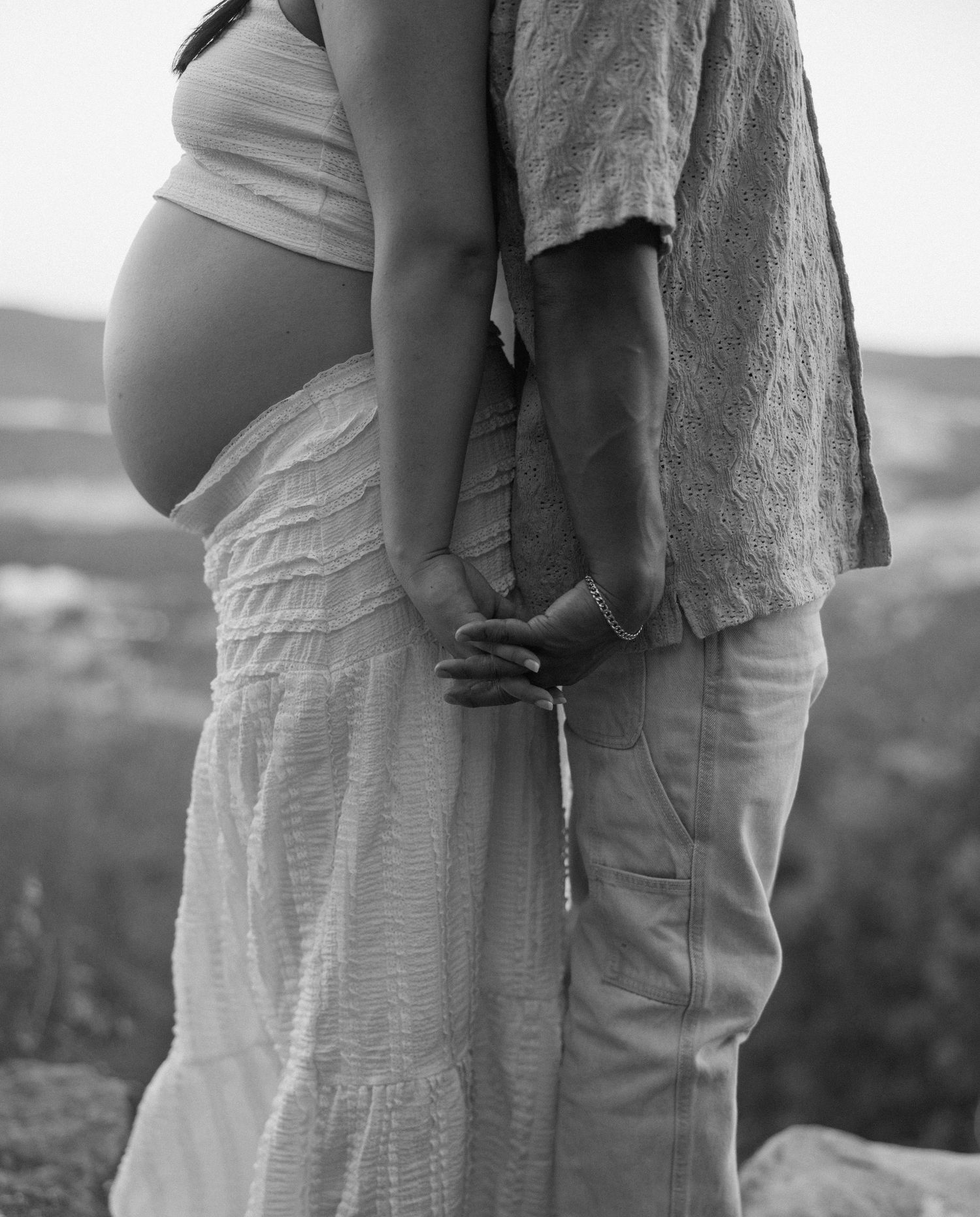 Black and white intimate maternity photo showing couple embracing outdoors with pregnant belly visible.