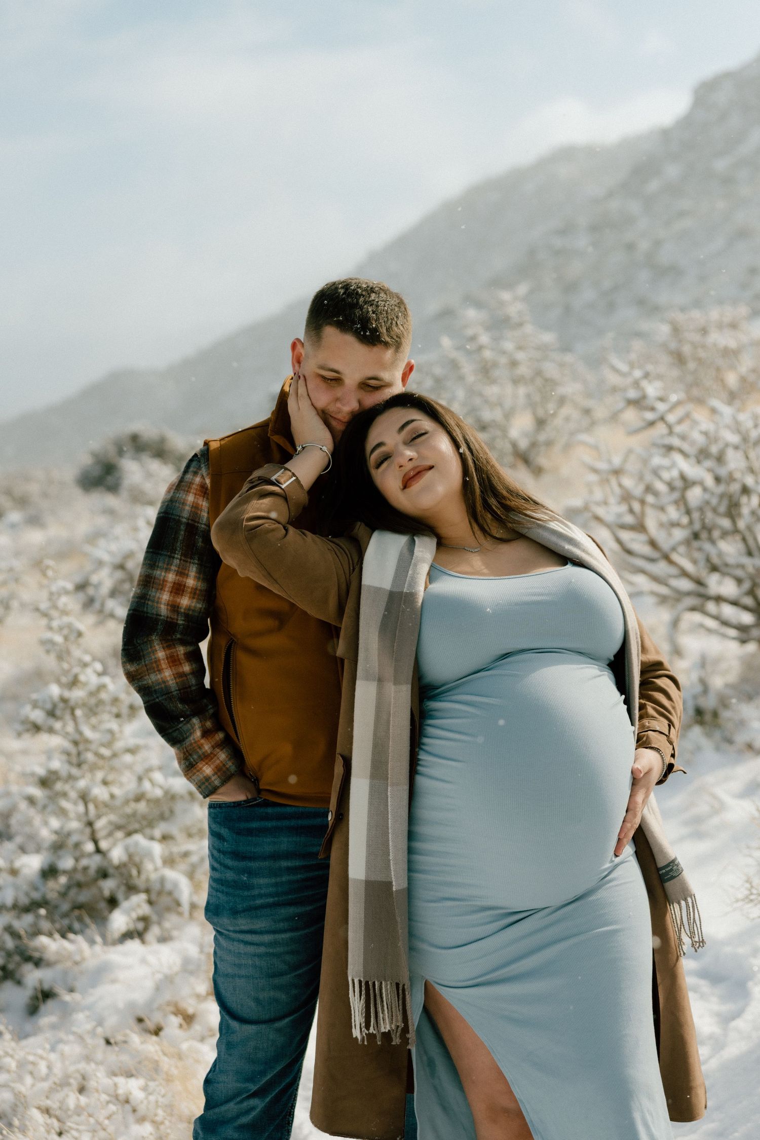 Maternity couple embraces in a snowy mountain landscape wearing winter clothing and a long scarf. Editorial maternity poses