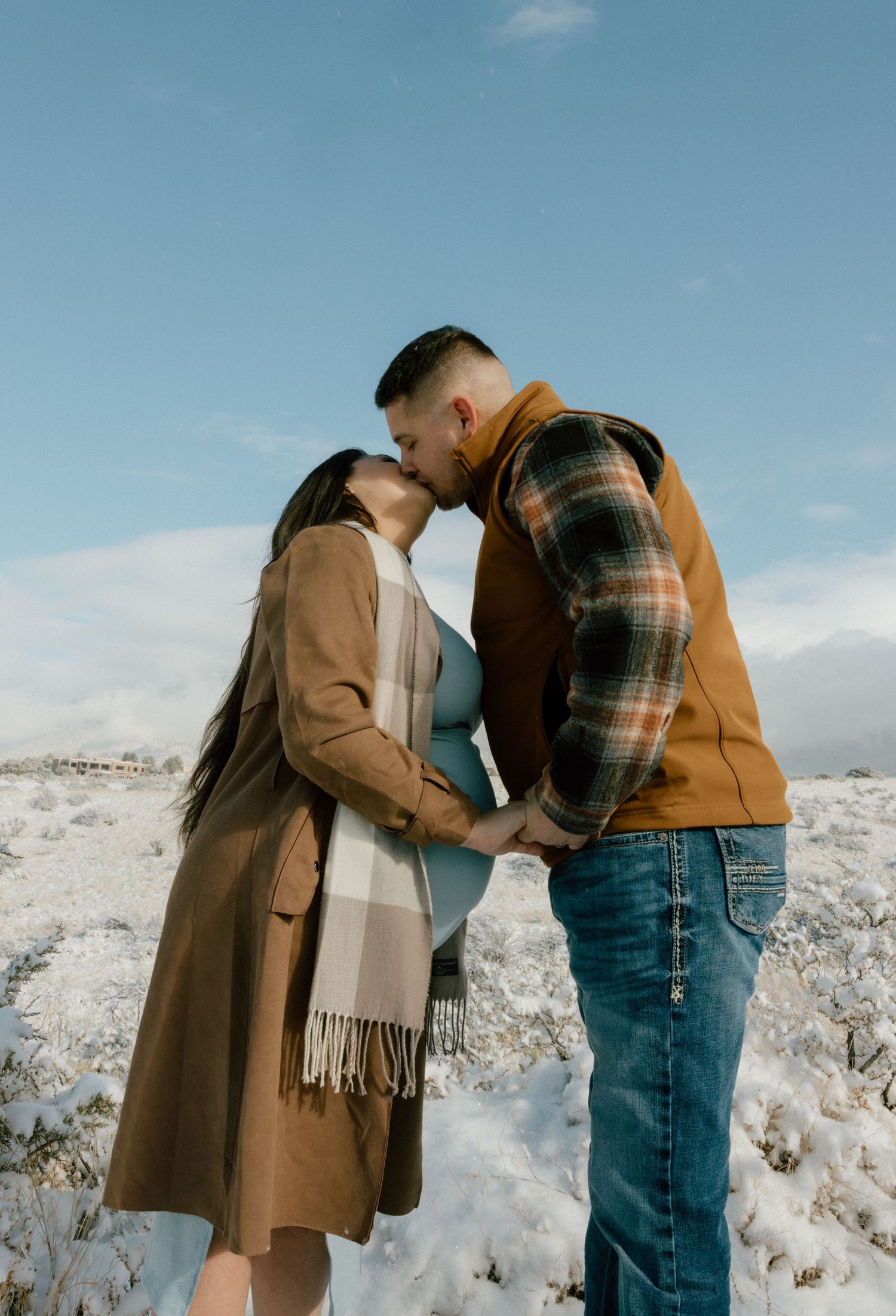 A couple shares intimate moments in a snowy winter landscape while wearing warm coats and casual attire.