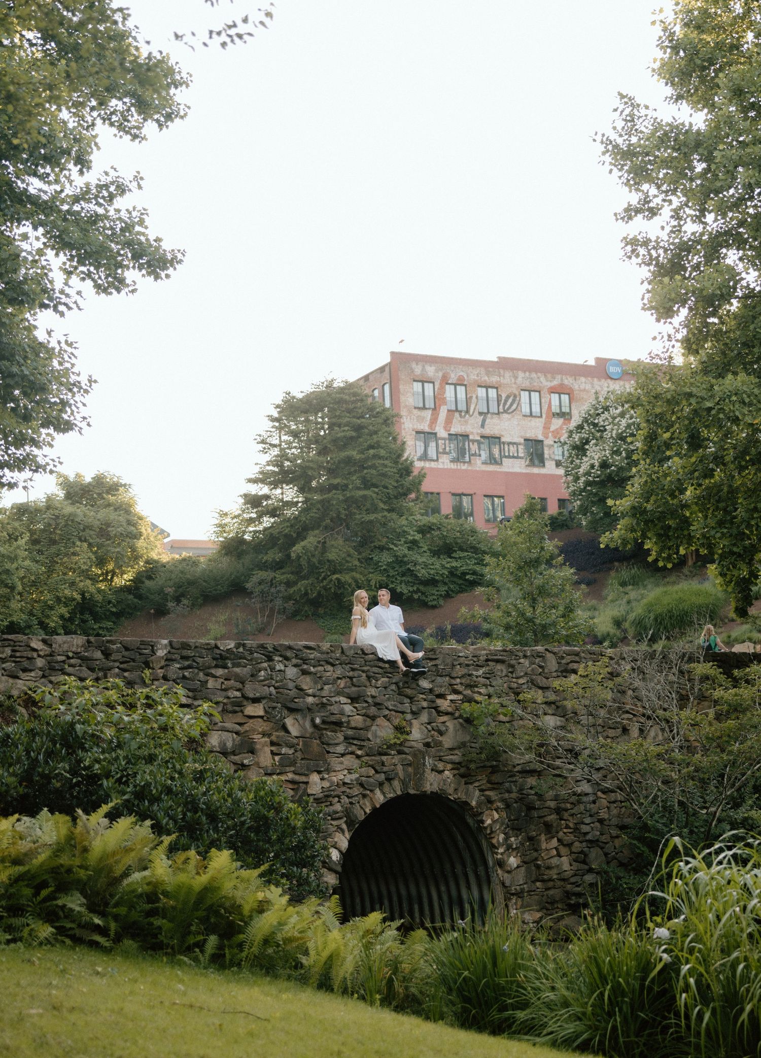 A stone wall with an arched opening winds through a lush garden landscape with trees and buildings in the background.