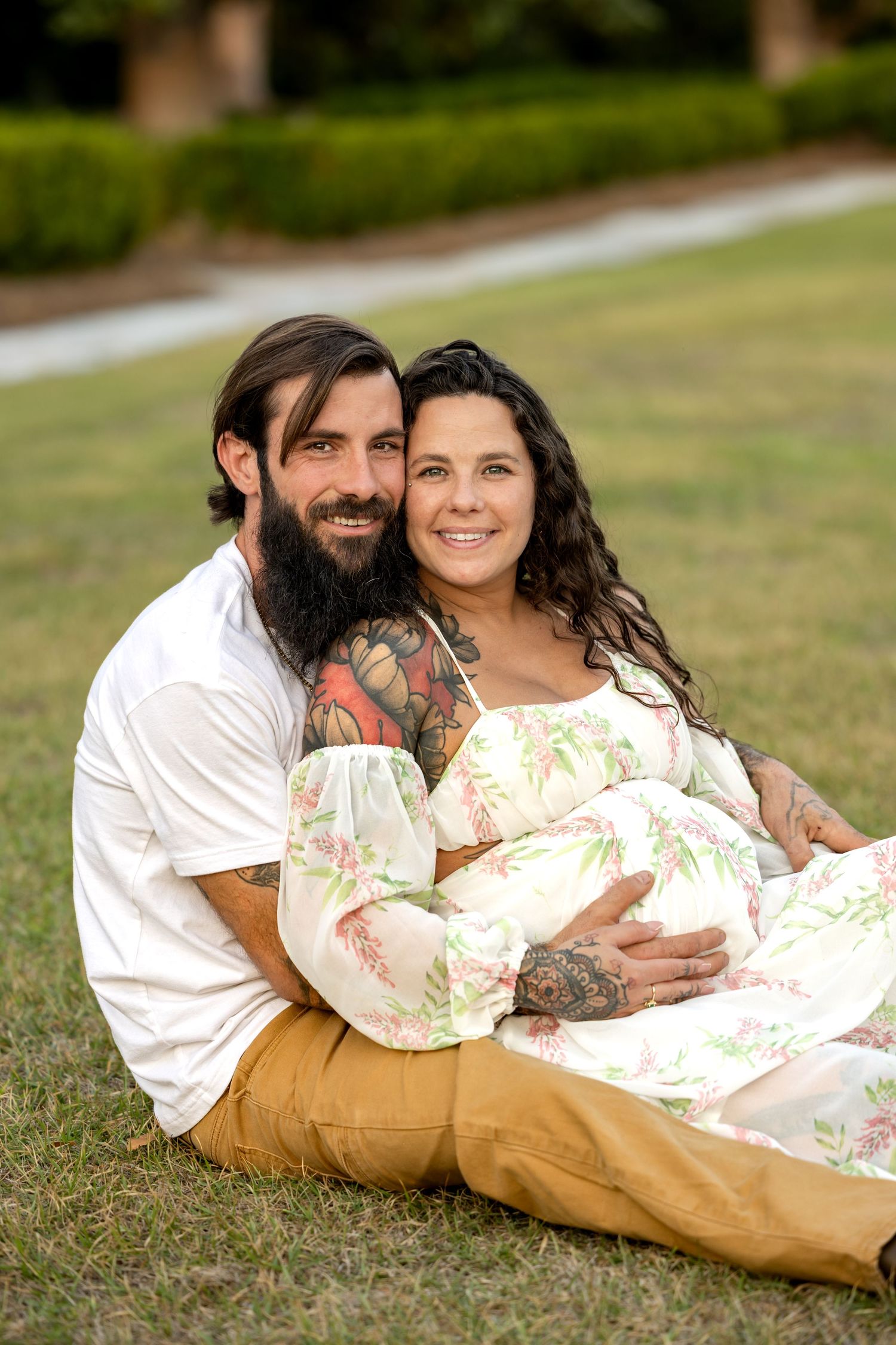 Tattooed couple sitting on grass, pregnant woman in floral dress cradled by bearded man, outdoor maternity photo session.
