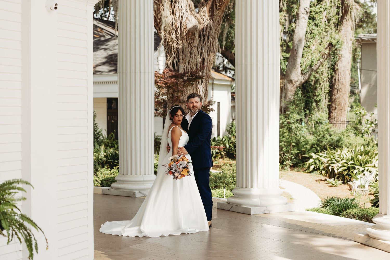 Elegant wedding portrait with white columns and lush greenery in the background.