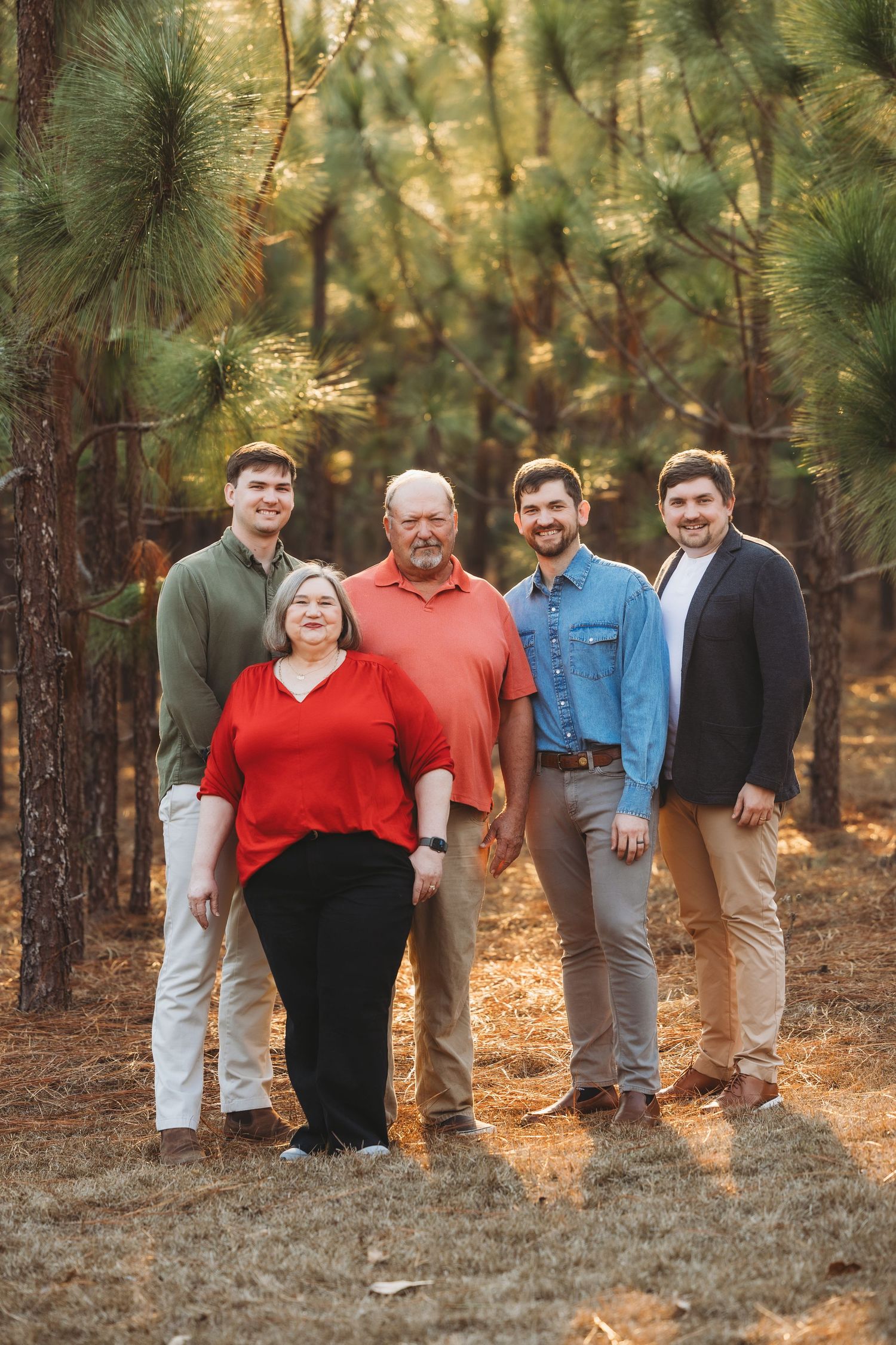 Family of five smiling together in a pine forest, wearing casual attire in warm autumn light.