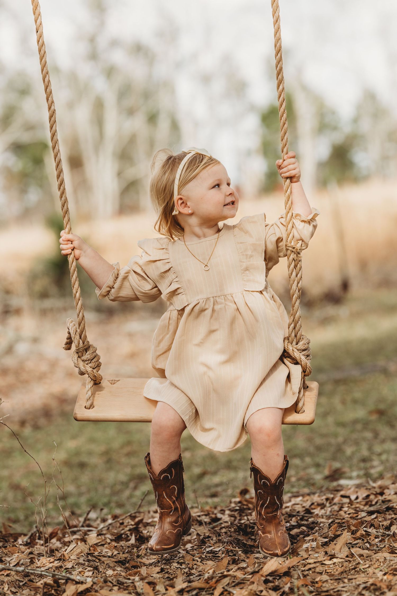 Young child in beige dress and boots enjoying a wooden rope swing outdoors.