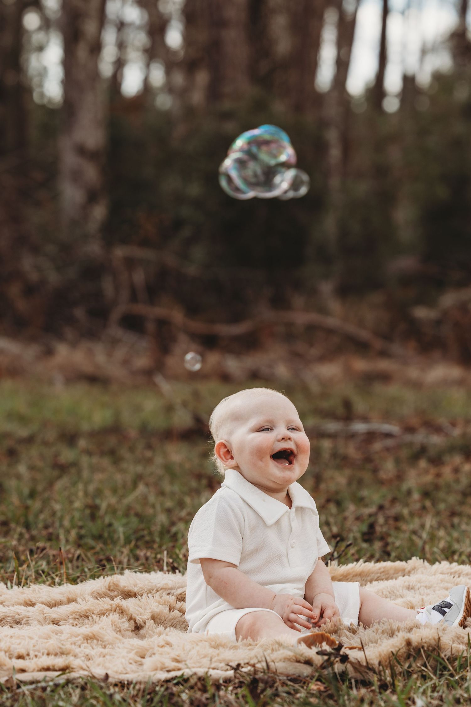 Baby in white outfit lying on blanket outdoors with soap bubbles floating above.
