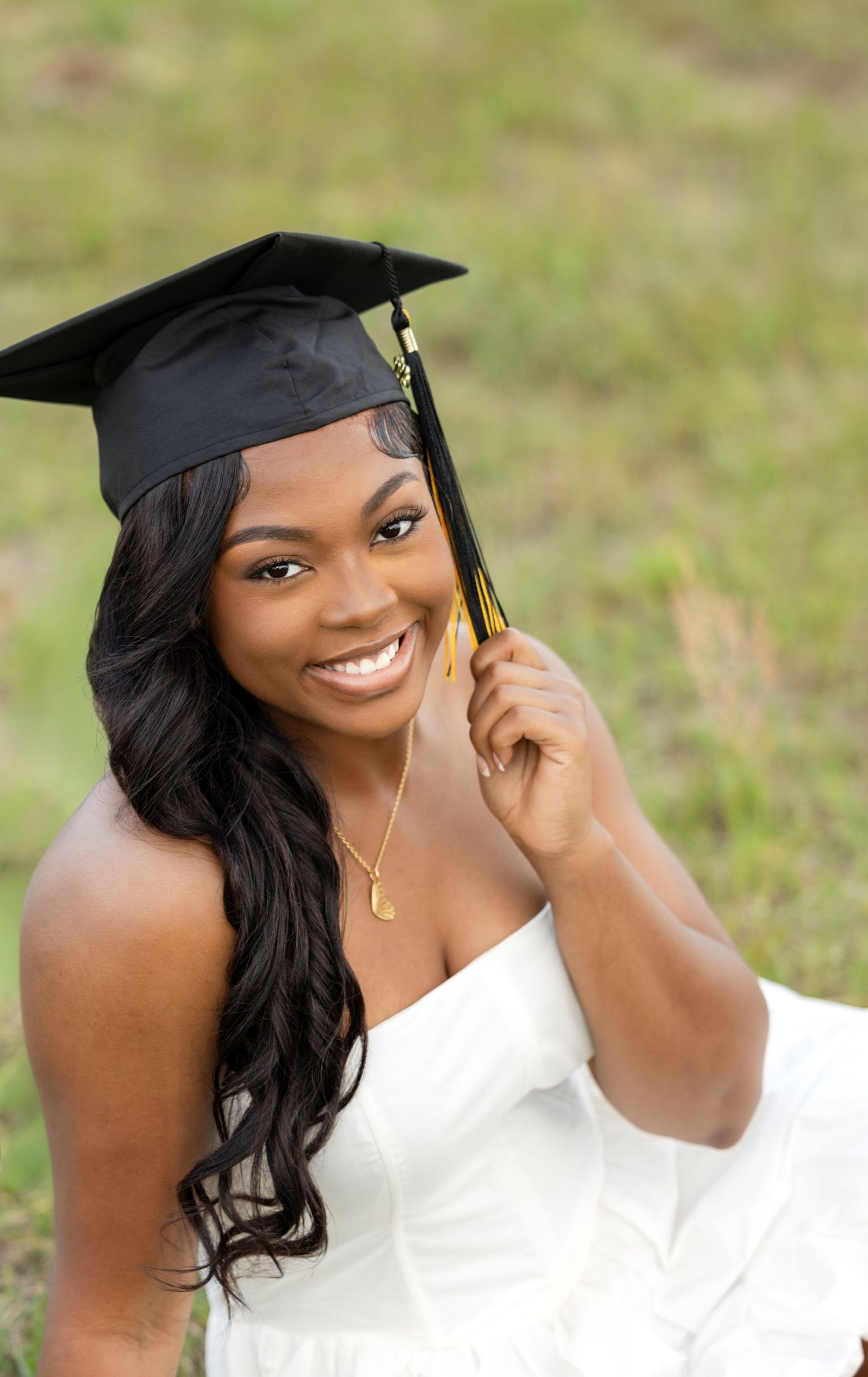 Smiling young woman in white dress and black graduation cap holding gold tassel outdoors.
