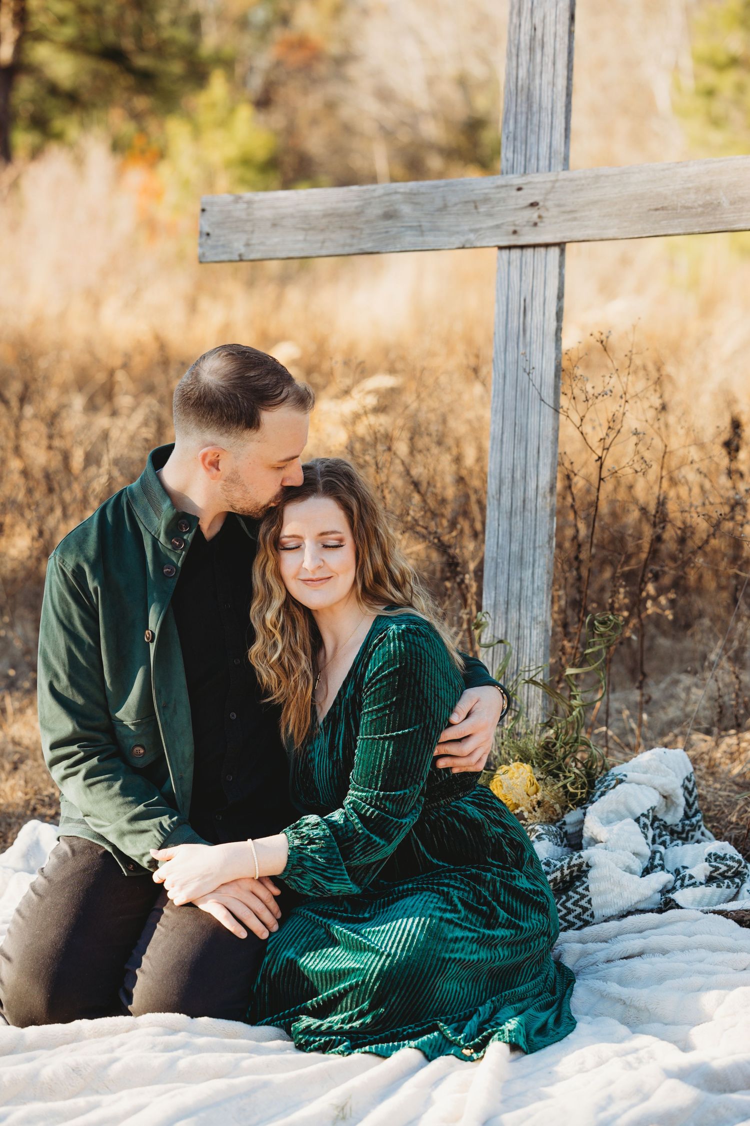 Couple in green velvet outfits sitting together near a wooden cross in an autumn field.