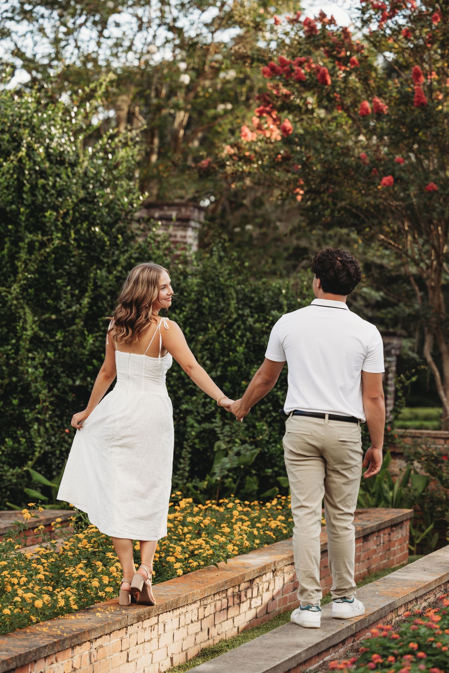 A couple walking hand in hand along a garden path with red flowers and greenery in the background.