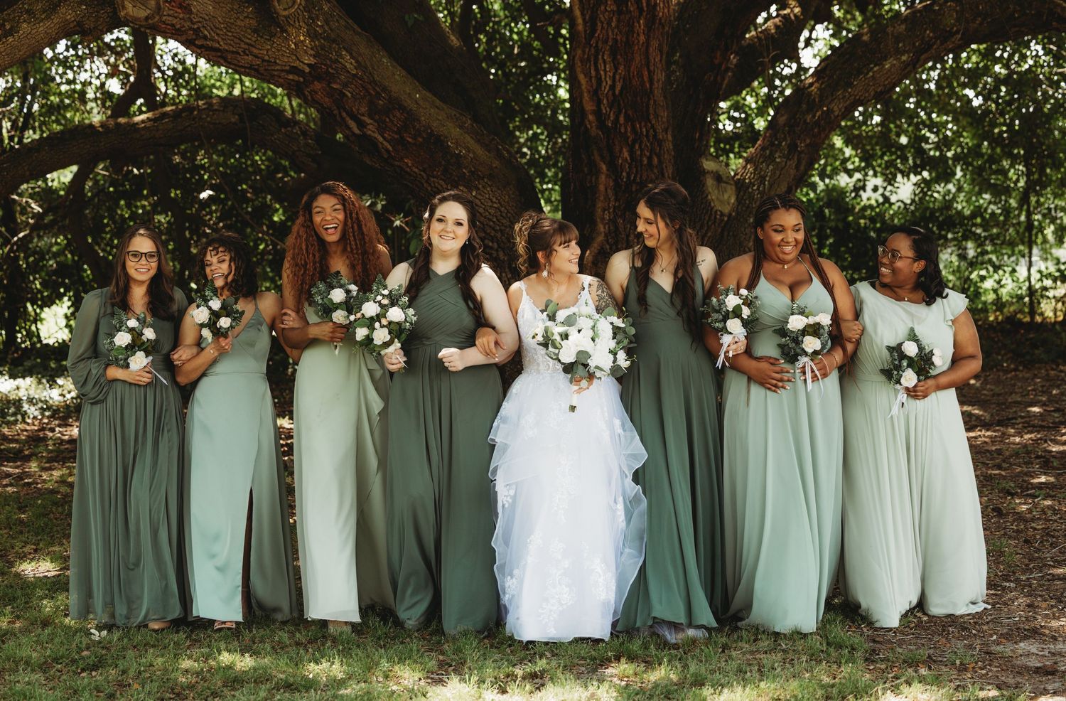A wedding party in sage green dresses poses together under a large tree.