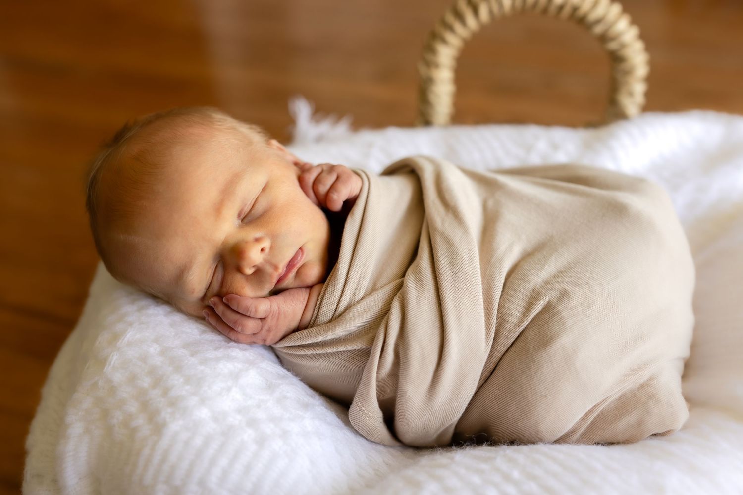 Peaceful newborn baby swaddled in a soft beige wrap, sleeping in a white wicker basket on a white blanket.