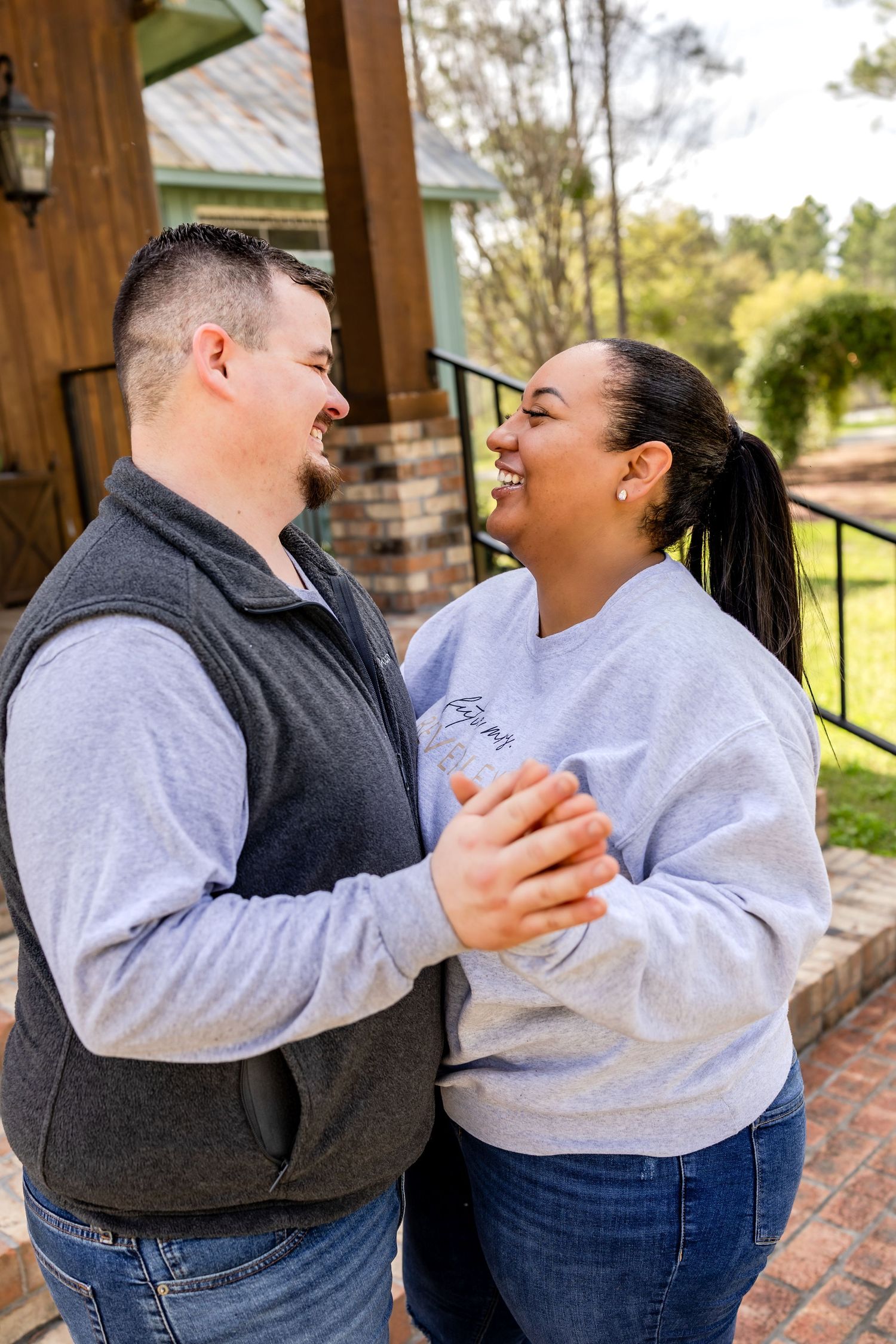 A smiling couple holds hands and gazes at each other outdoors near a wooden building surrounded by trees.