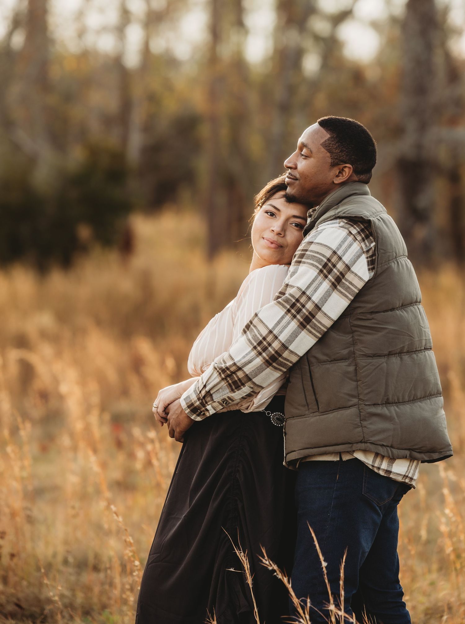 Couple embracing in golden autumn field, woman leaning back into mans arms, surrounded by tall grass and trees.