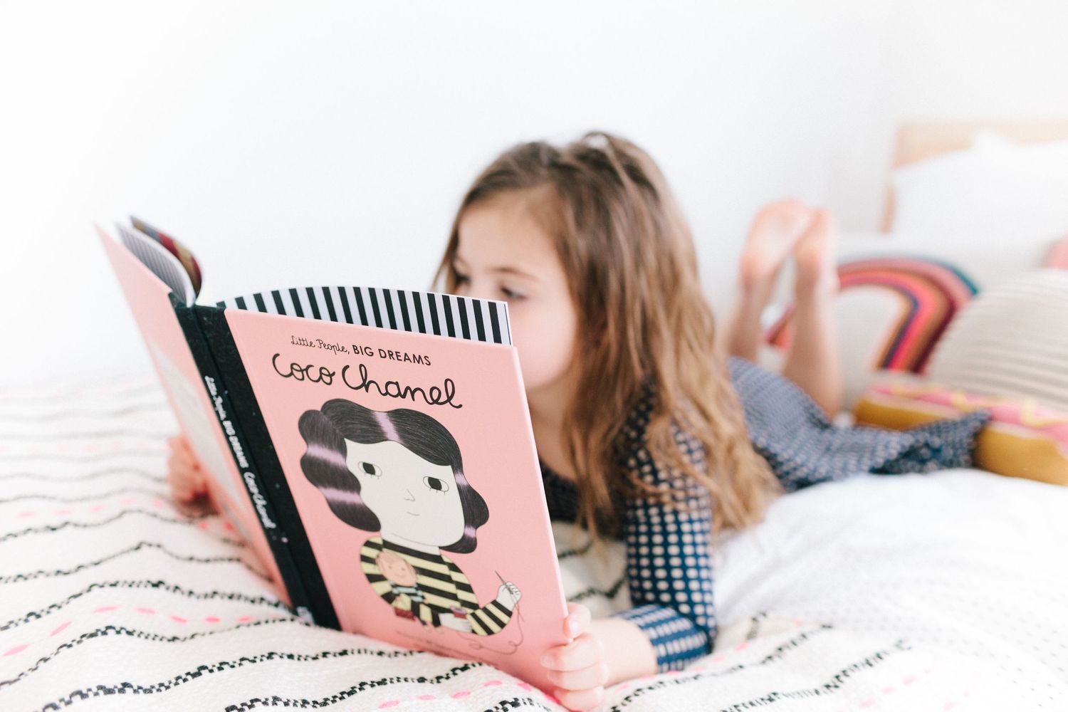 little girl reading "little people, big dreams: coco chanel" while laying on her stomach on a bed