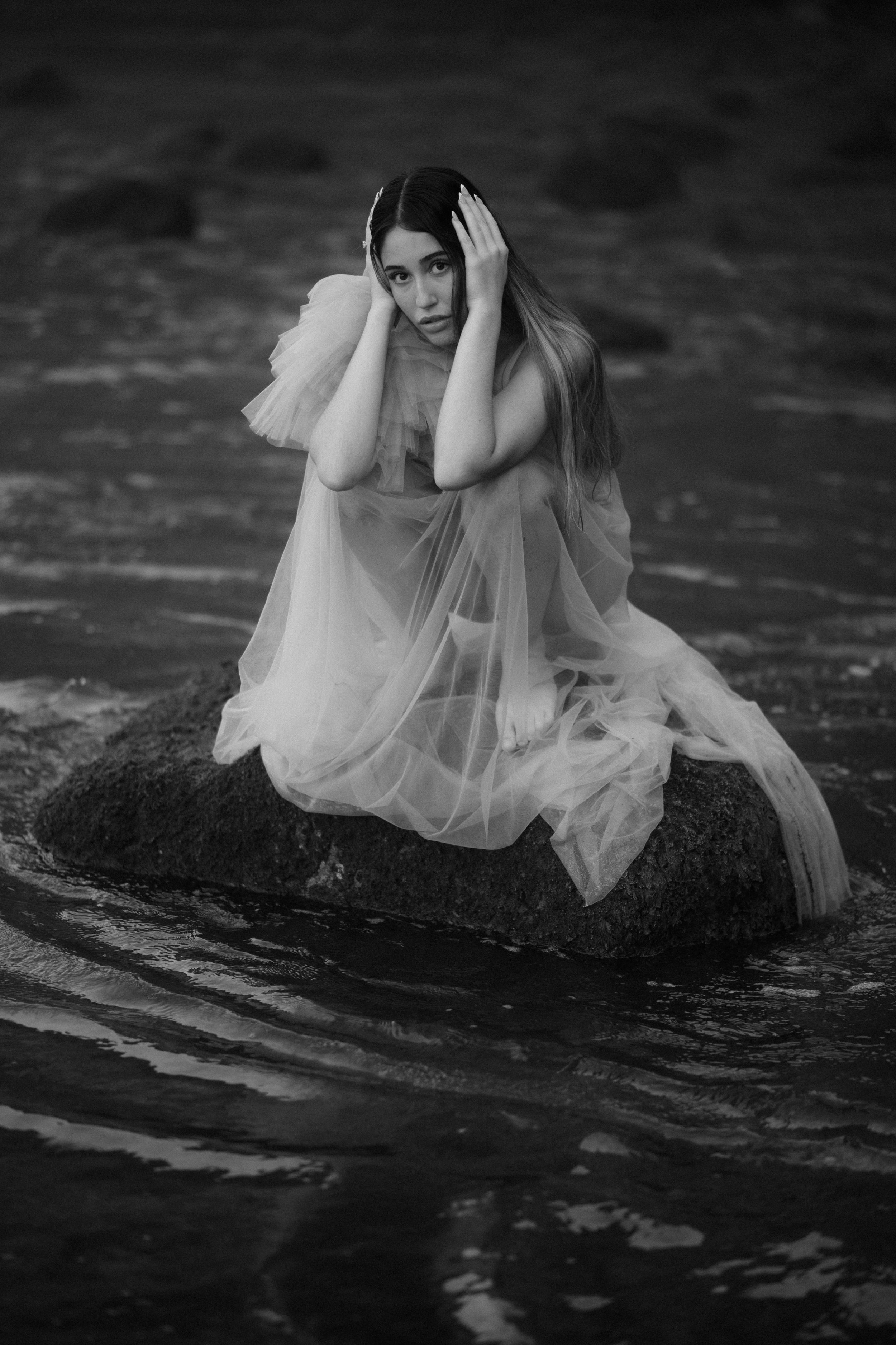 A figure in flowing white fabric sits gracefully in dark rippling water creating a dramatic black and white artistic scene.