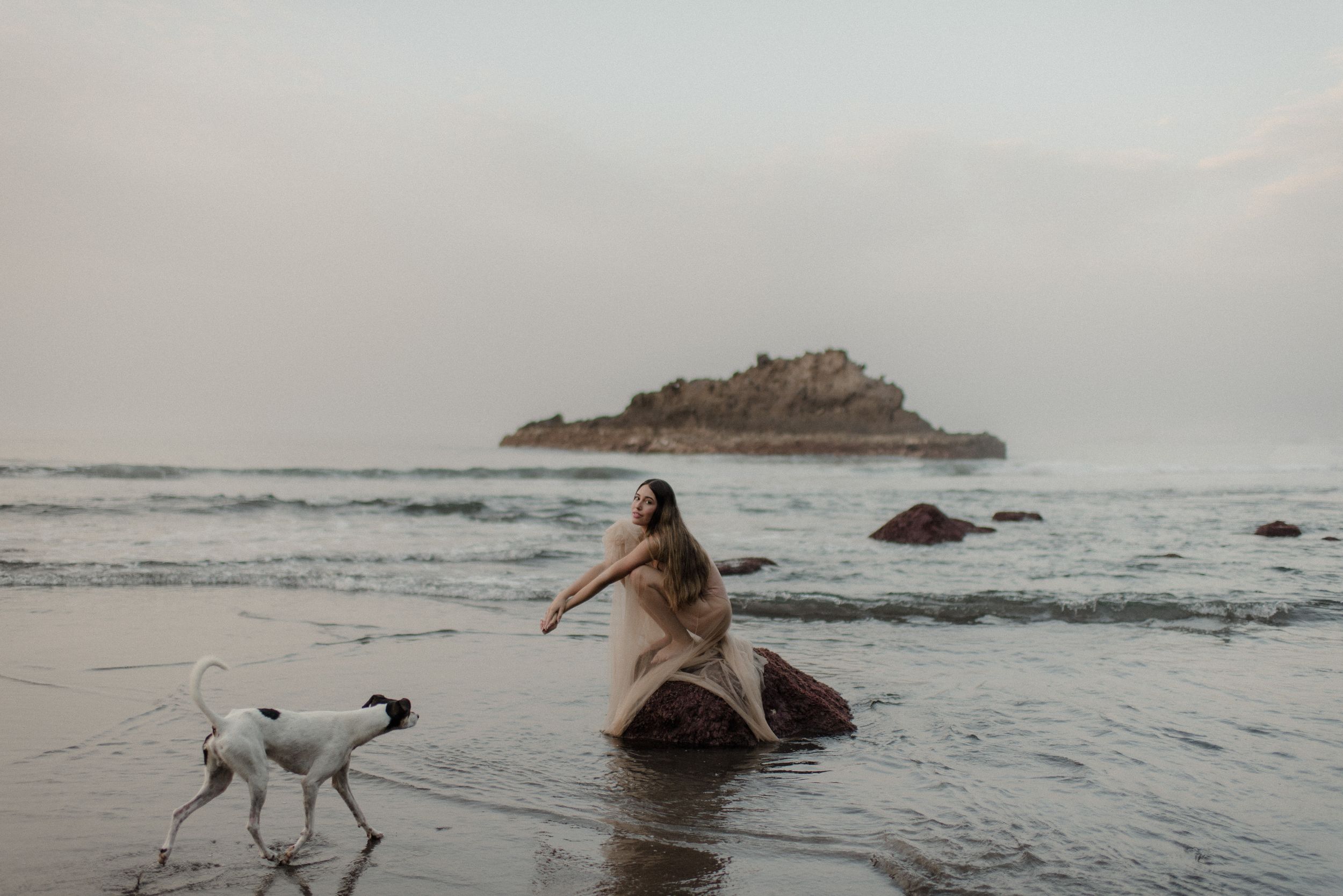 Scenic coastal beach landscape with dark rocks and small islands visible in the misty ocean waters at sunset.
