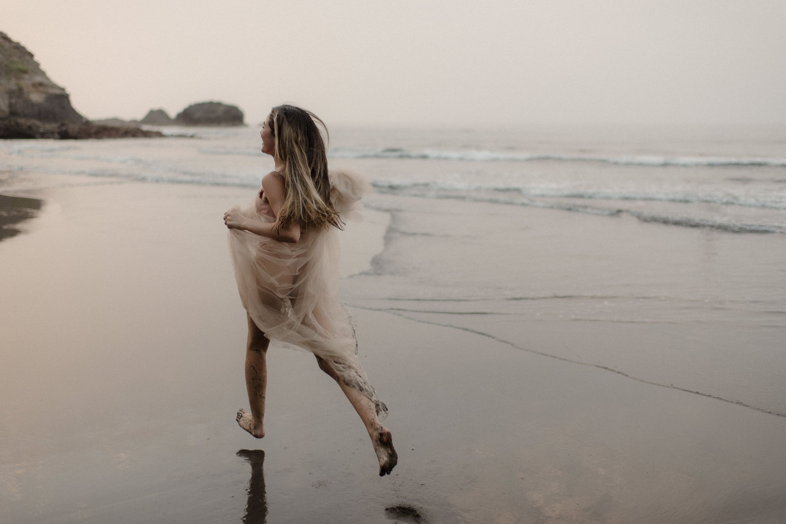 A figure in flowing white clothing dances gracefully along a misty beach shoreline.