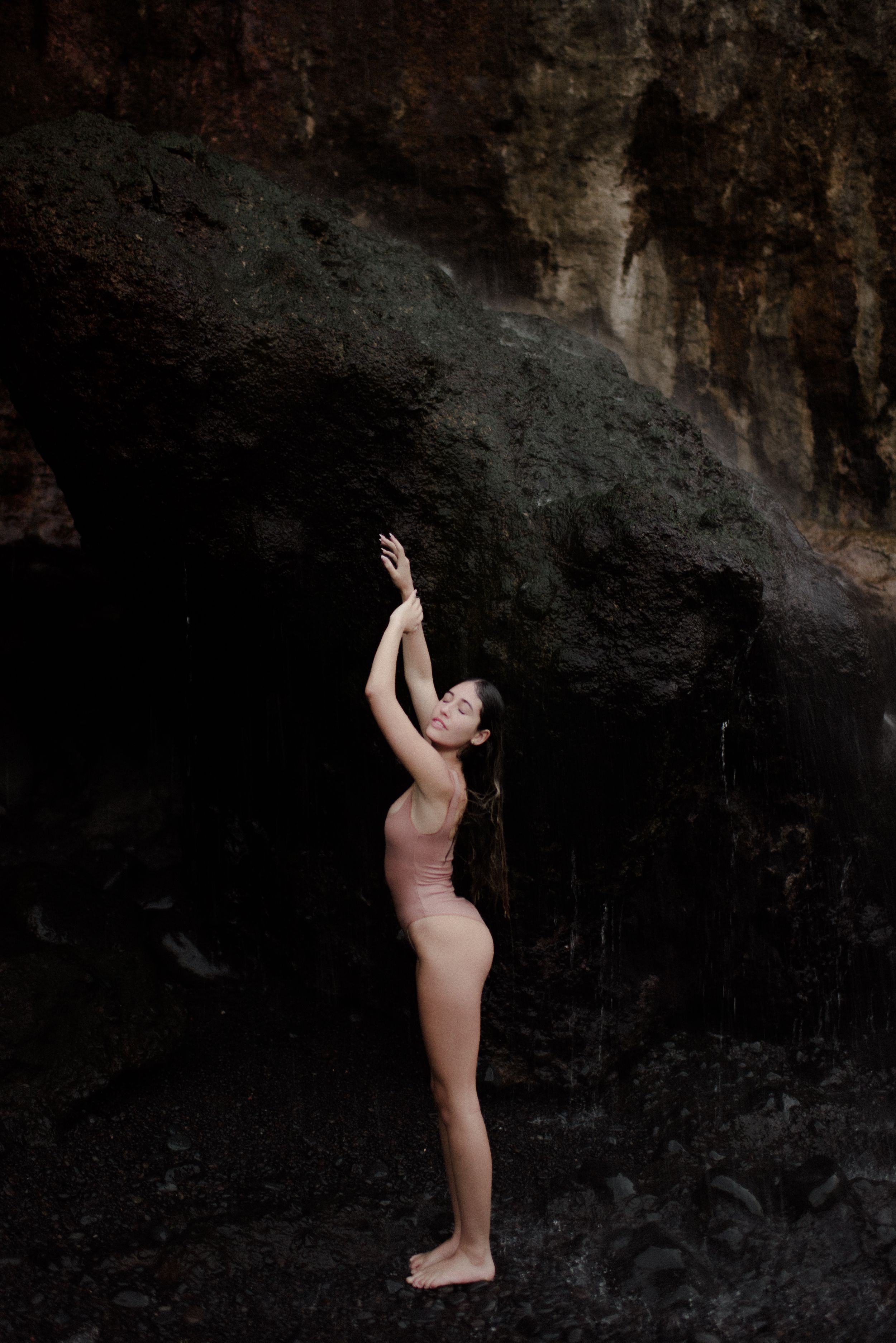 A figure poses gracefully against a dark rocky cliff backdrop near the ocean.