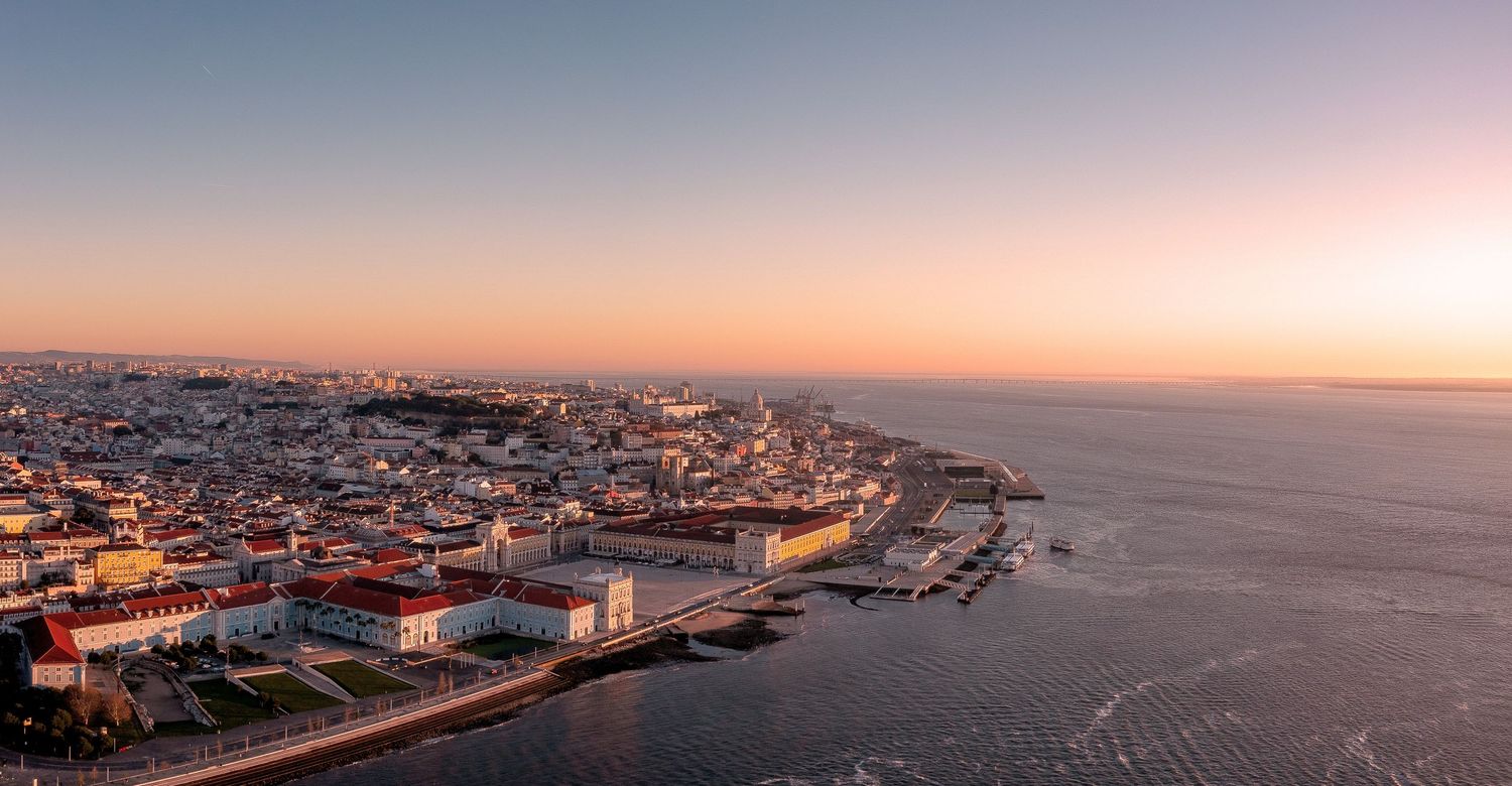 Panoramic aerial view of Lisbon's historic waterfront and Tagus River during a colorful sunset.