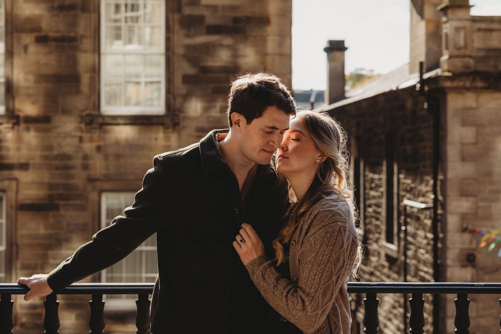 A romantic couple embraces on a balcony against a rustic stone building backdrop on a sunny afternoon.