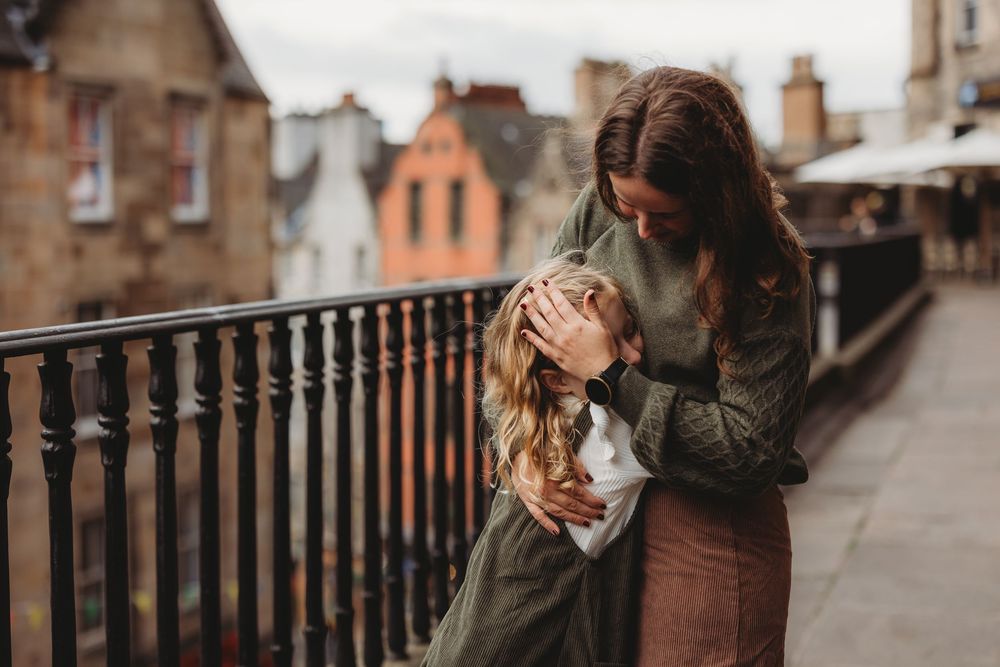 Two people share an intimate embrace on a balcony overlooking historic stone buildings in Edinburgh.