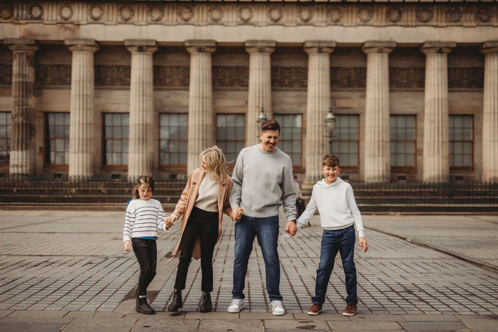 A family walks together holding hands along a cobblestone plaza near a historic building with columns.