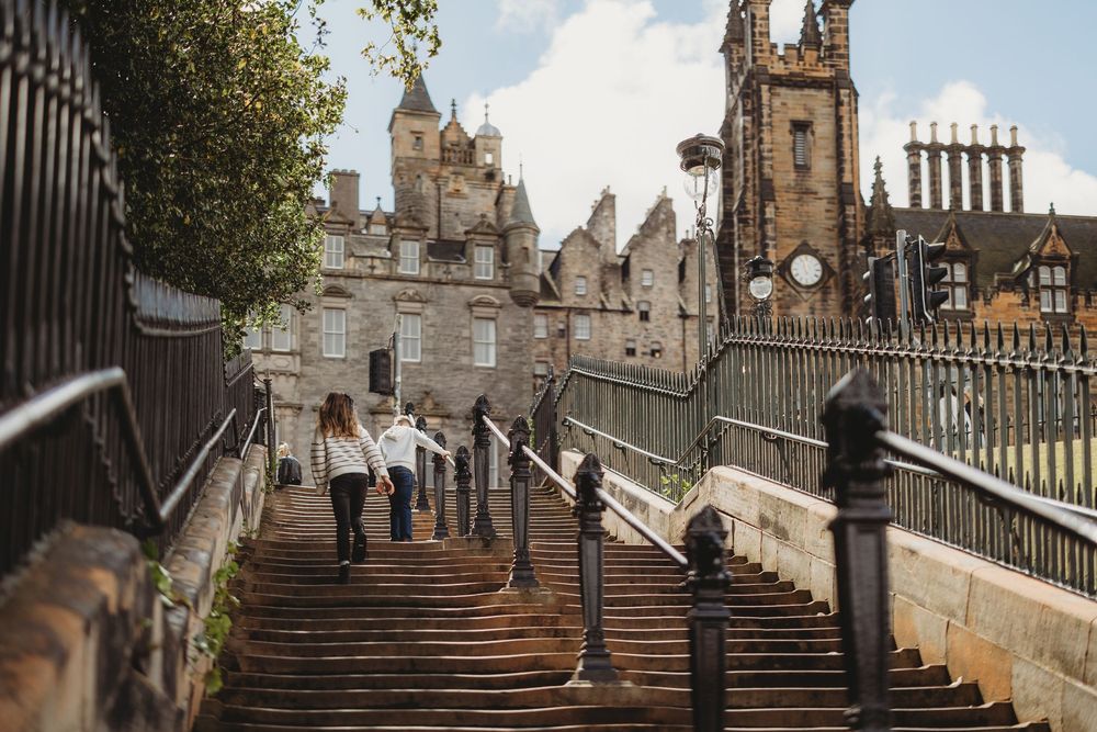 Historic stone staircase with ornate iron railings leads up to Edinburgh's Old Town buildings and church spires.