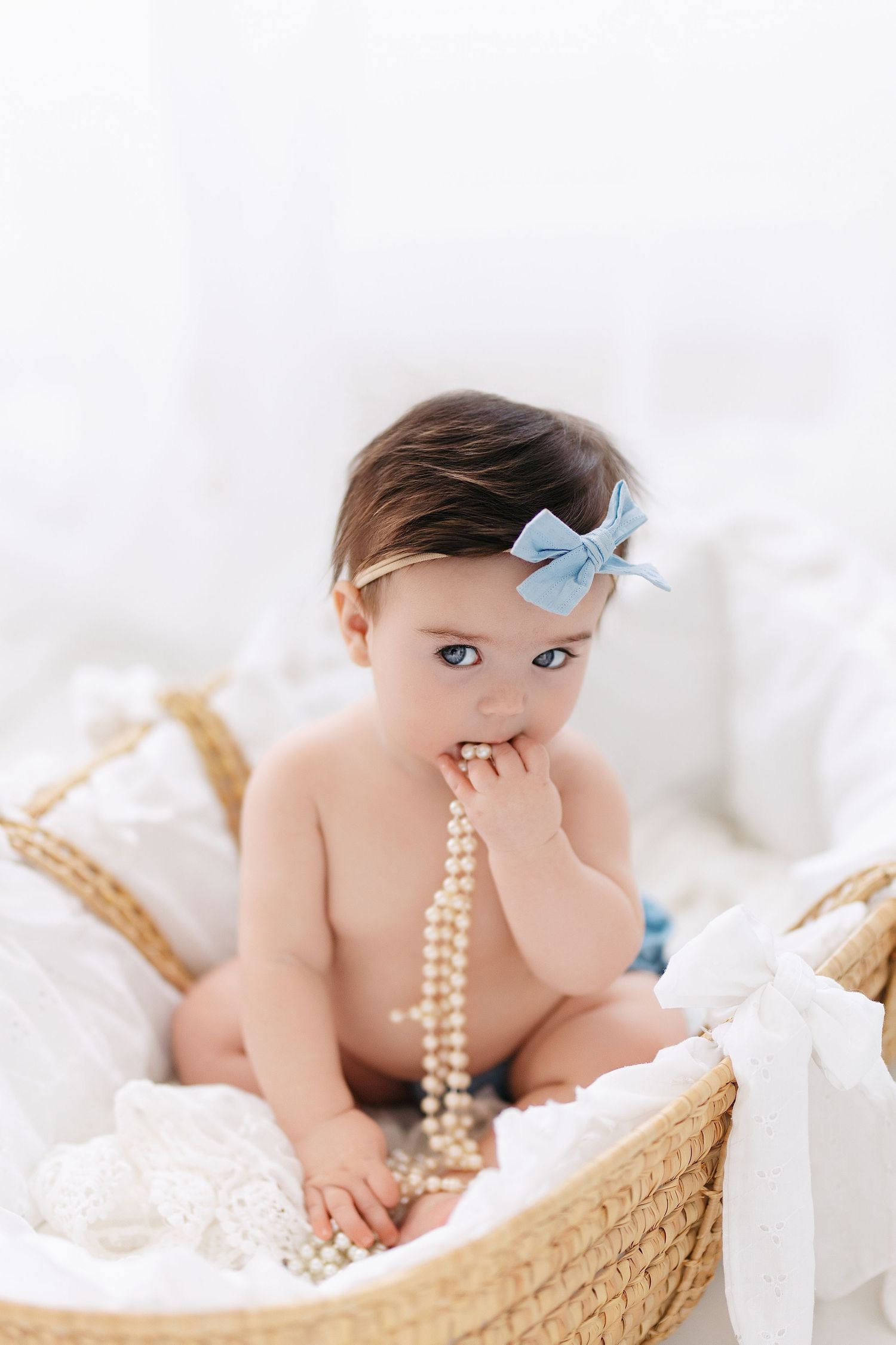 Baby wearing blue bow and pearls sits in woven basket with white linens during portrait session.